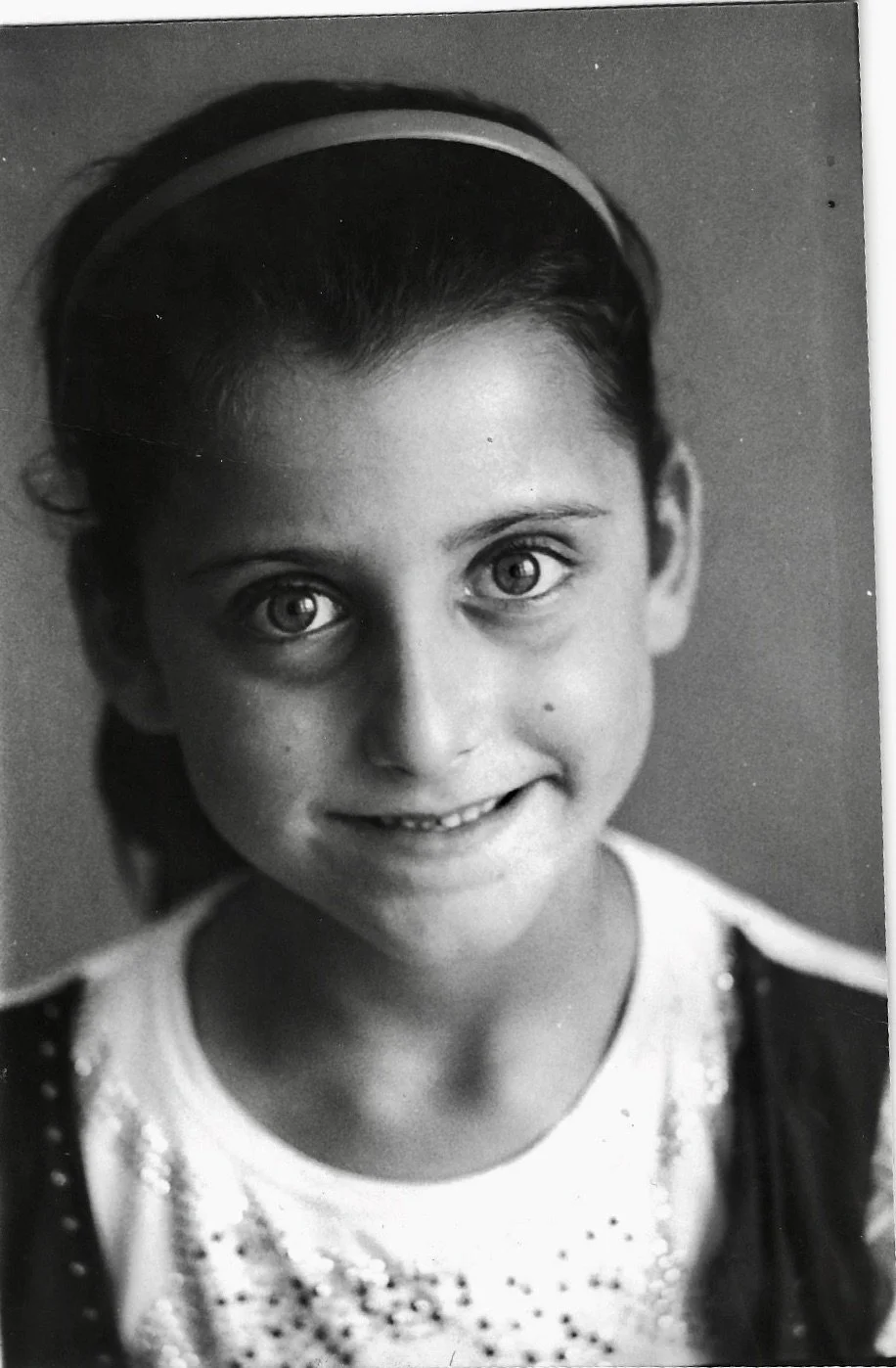 Black and white portrait of a young girl with large eyes and a bright smile, wearing a headband and a sparkly top.
