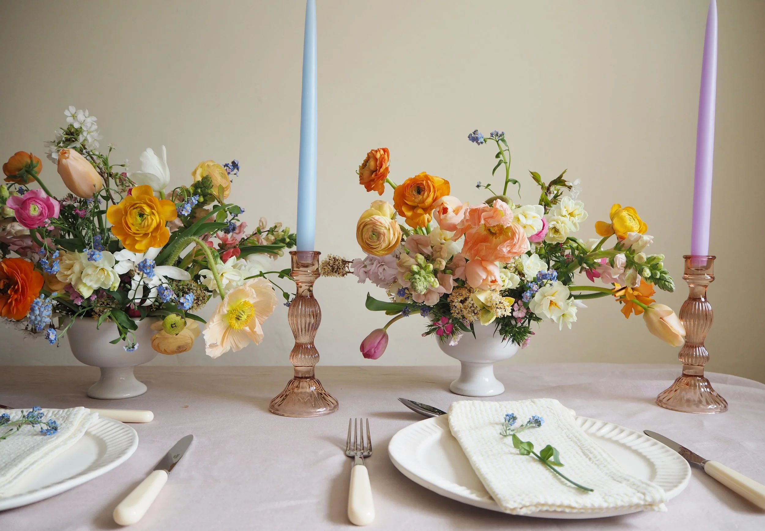 a wedding reception table with two white footed bowls full of spring flowers, glass candlesticks with pale blue and lilac dinner candles