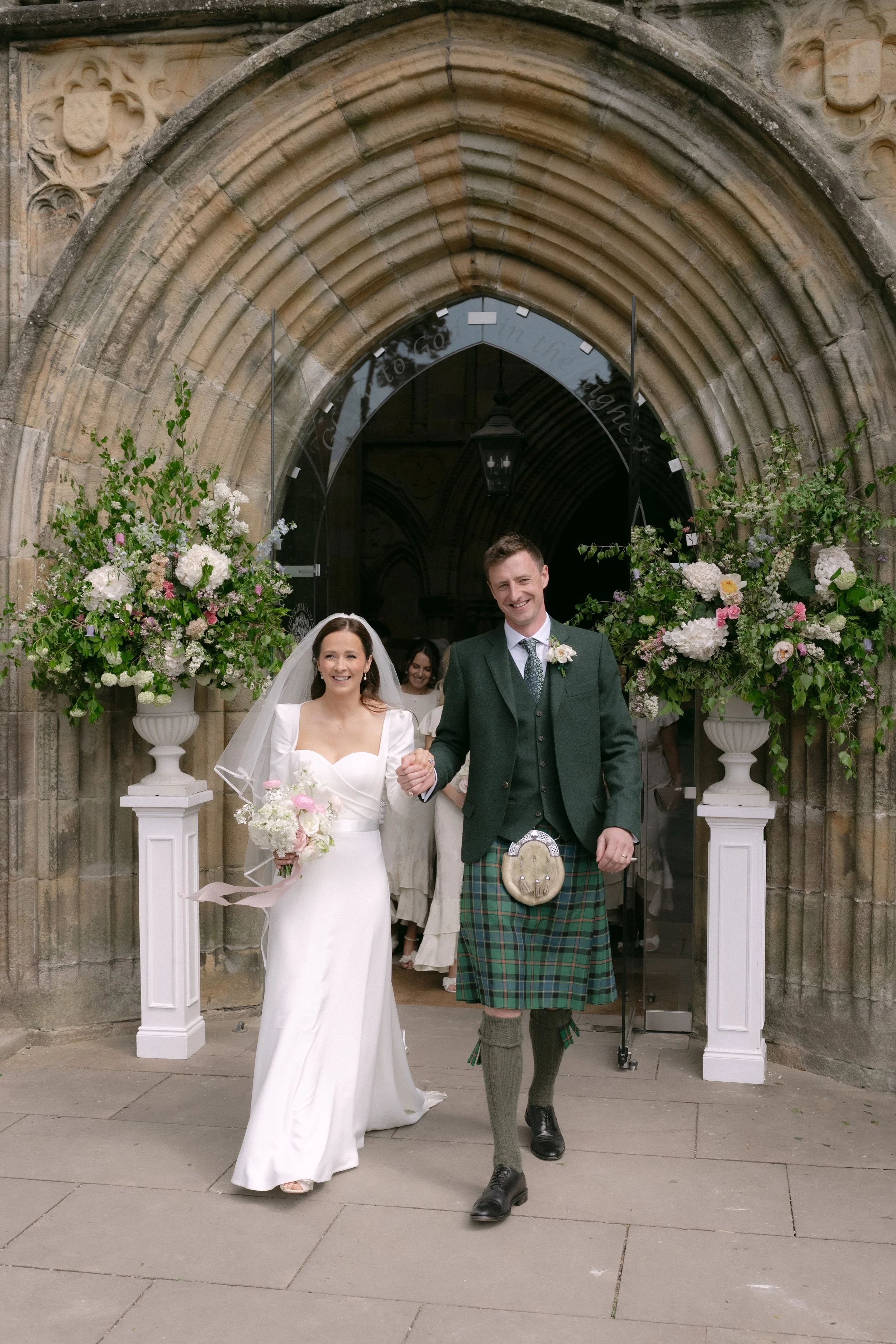 A newlywed couple walking out of a church after their wedding, holding hands and smiling. The bride wears a white wedding dress and veil, carrying a bouquet of flowers. The groom wears a green plaid kilt, jacket, and tie. Floral arrangements flank th