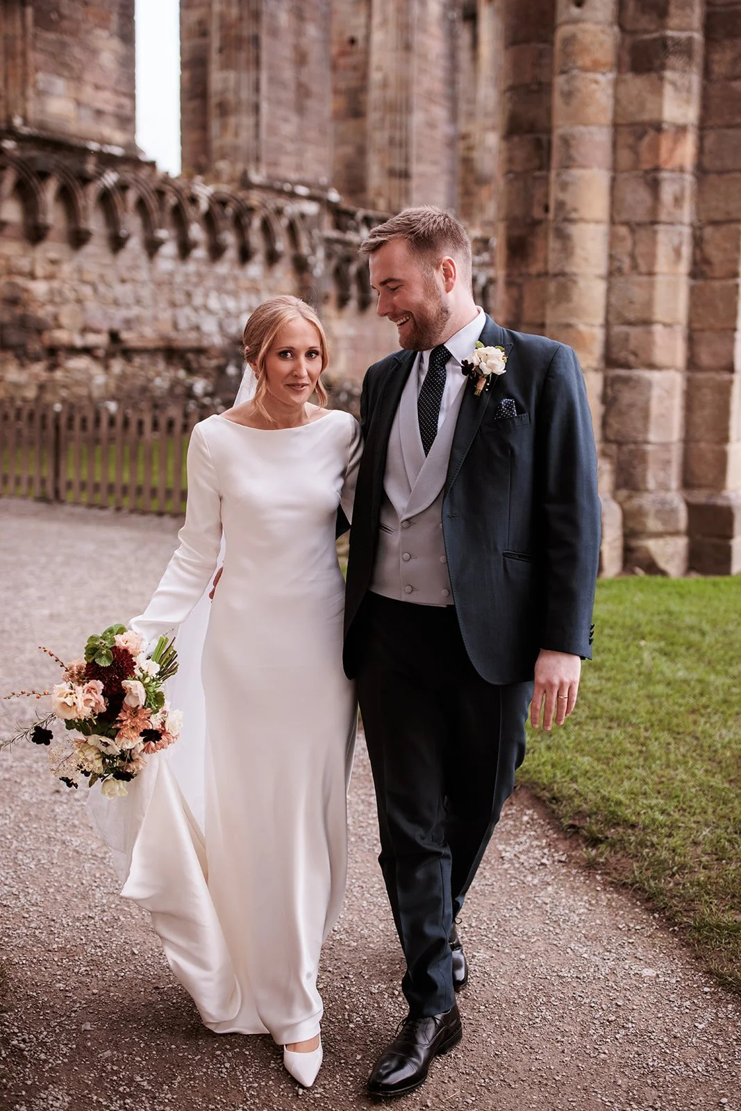 A bride and groom smiling and walking outdoors near a stone building, with the bride holding a bouquet of flowers, dressed in a white wedding gown, and the groom wearing a dark suit with a boutonnière.