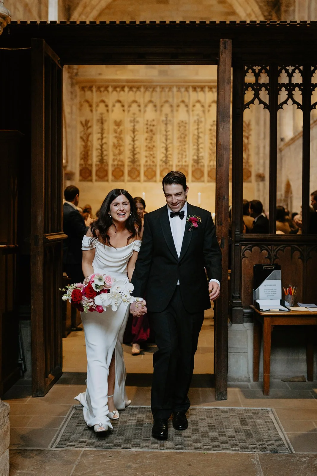 A bride and groom entering a wedding venue, holding hands and smiling, with guests in the background inside a decorated church or hall.