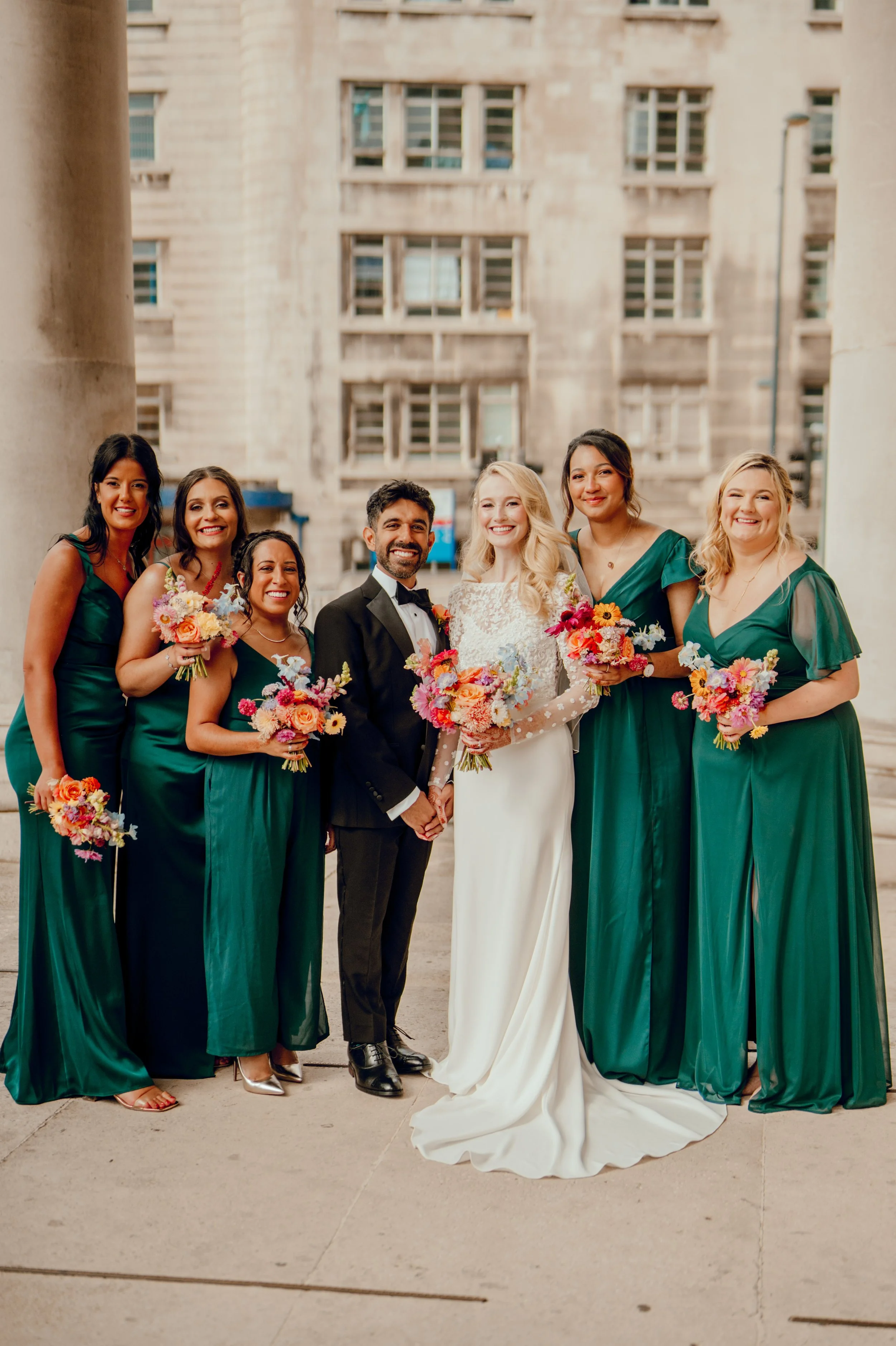Bride and groom with six bridesmaids standing outside a building, all holding bouquets of flowers.
