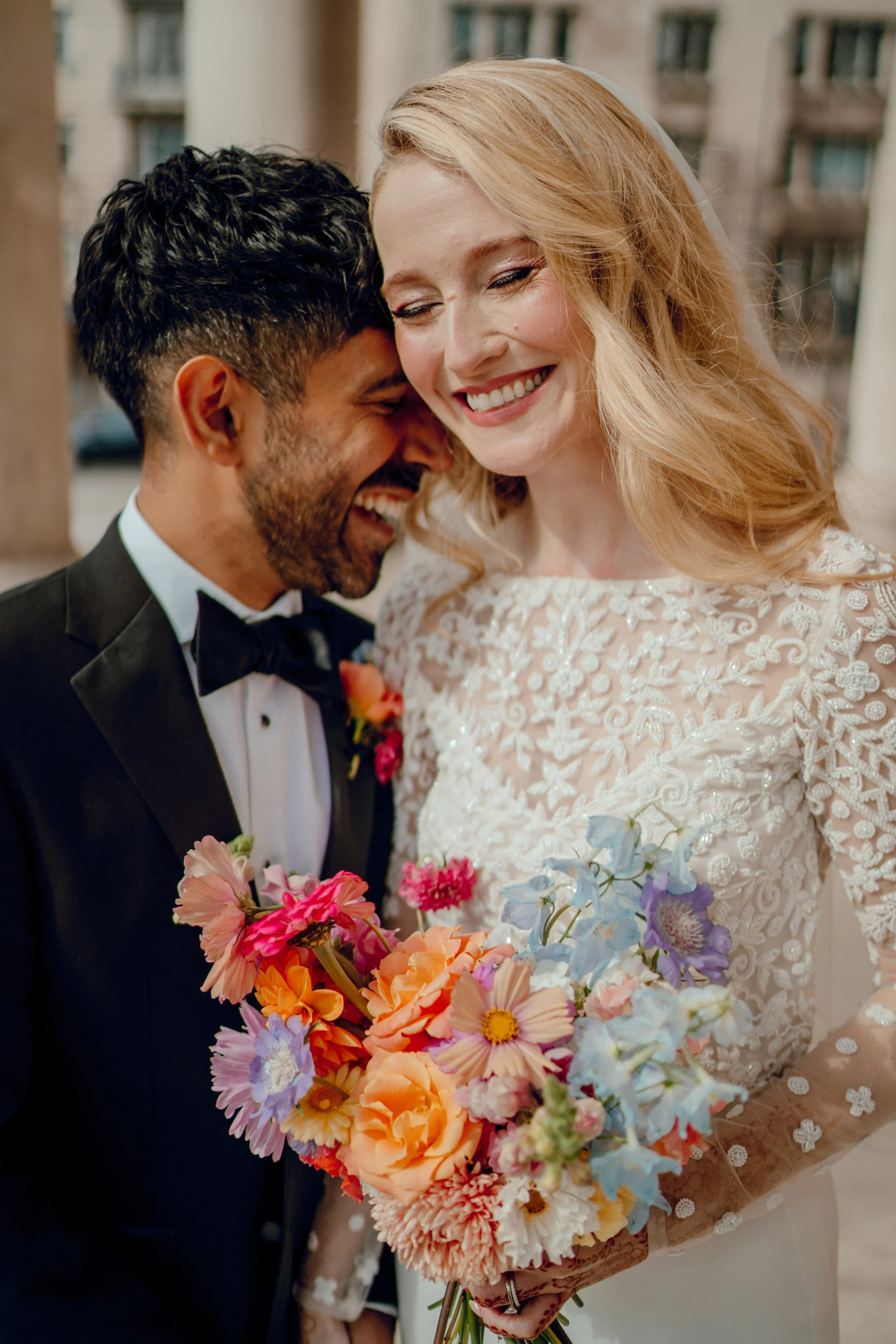 A happy couple dressed in wedding attire, the man in a tuxedo and the woman in a lace wedding dress, smiling and close together, holding a colorful bouquet of flowers.