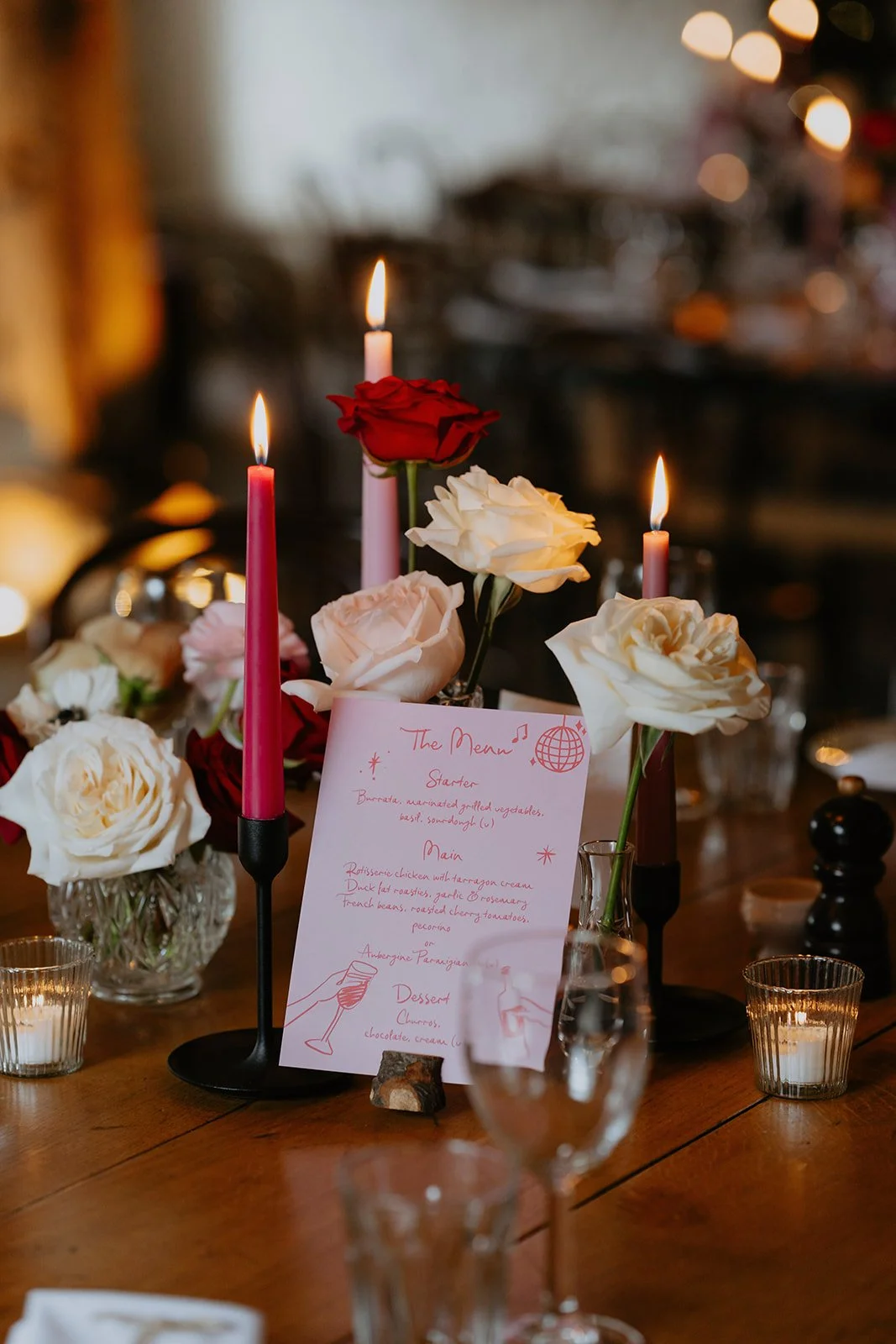 A candlelit table centerpiece featuring white and red roses, with a handwritten menu card, small candles, and a few wine glasses on a wooden table in a cozy, dimly lit setting.