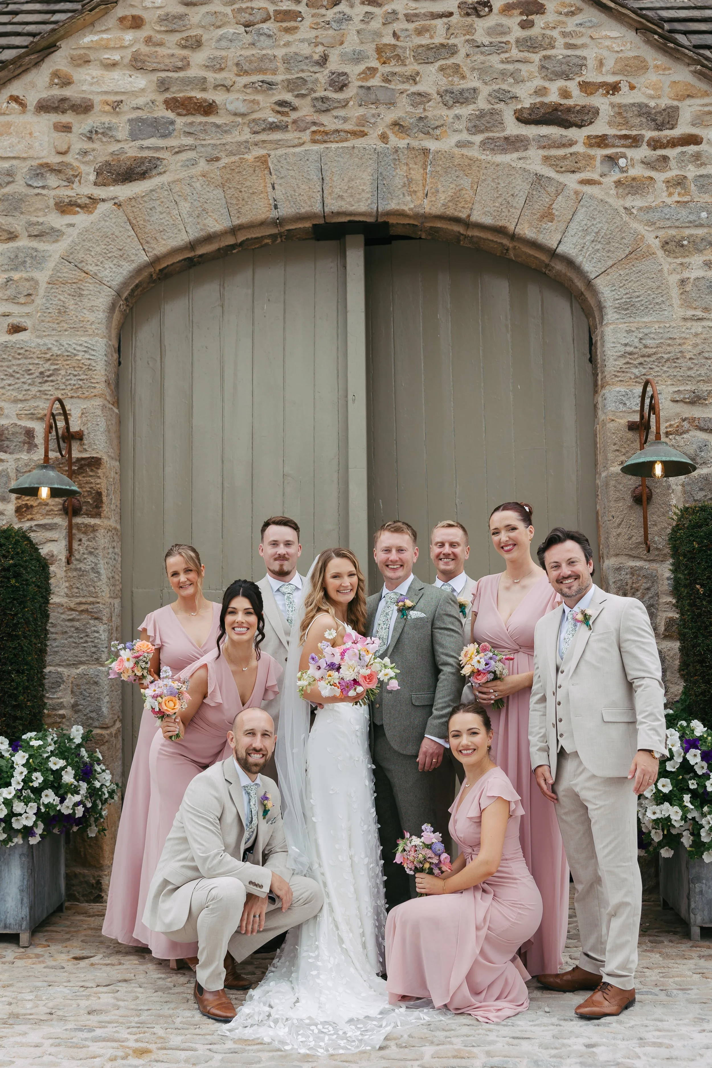 Just married couple with their groomsmen and bridesmaids outside the doors of the Tithe Barn, Bolton Abbey, Yorkshire. The bridesmaids are in pastel pink, flowers are pinks, whites and lilacs 