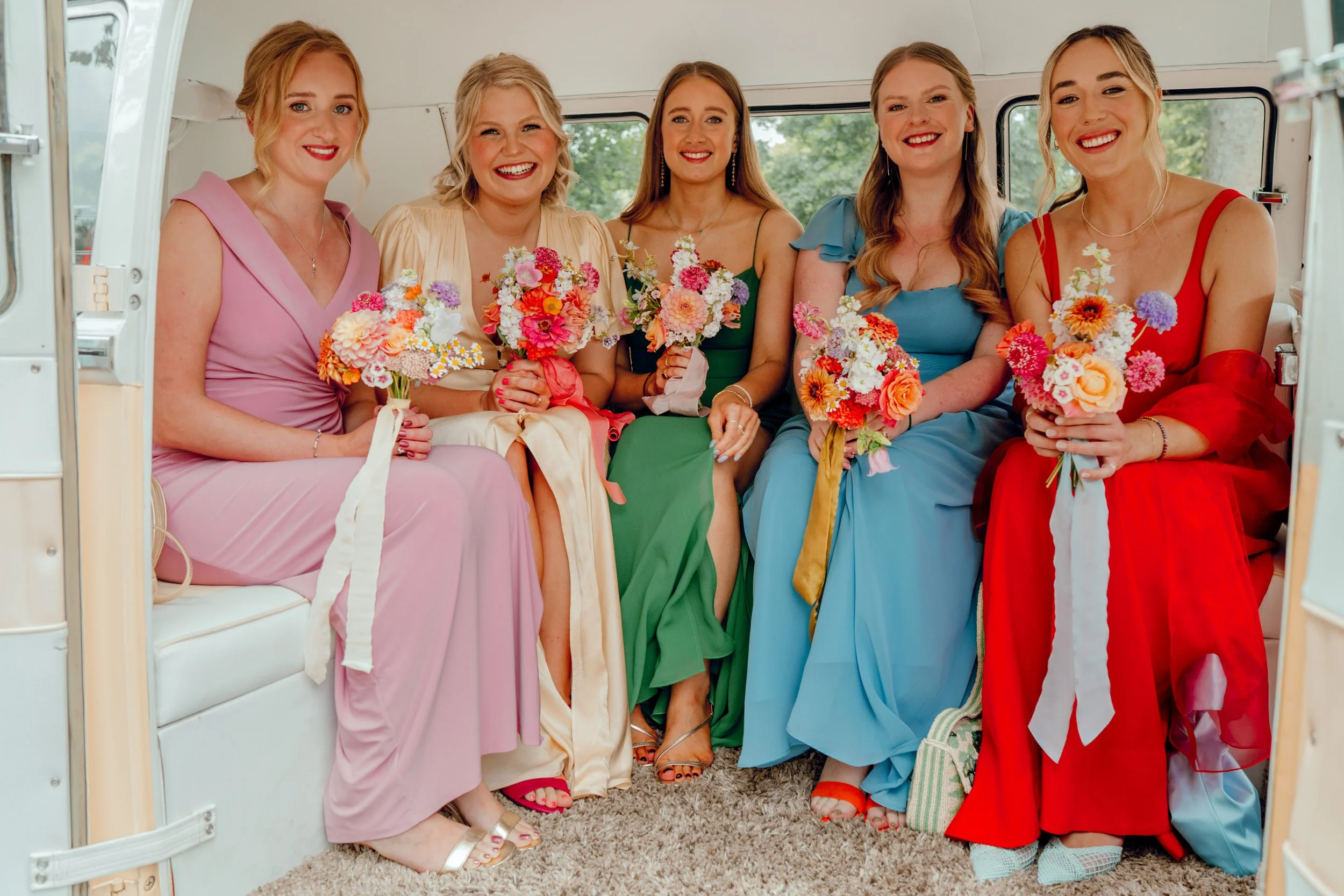 Bridesmaids in pink, green, blue, red and cream with brightly coloured bouquets sat in a vintage VW van