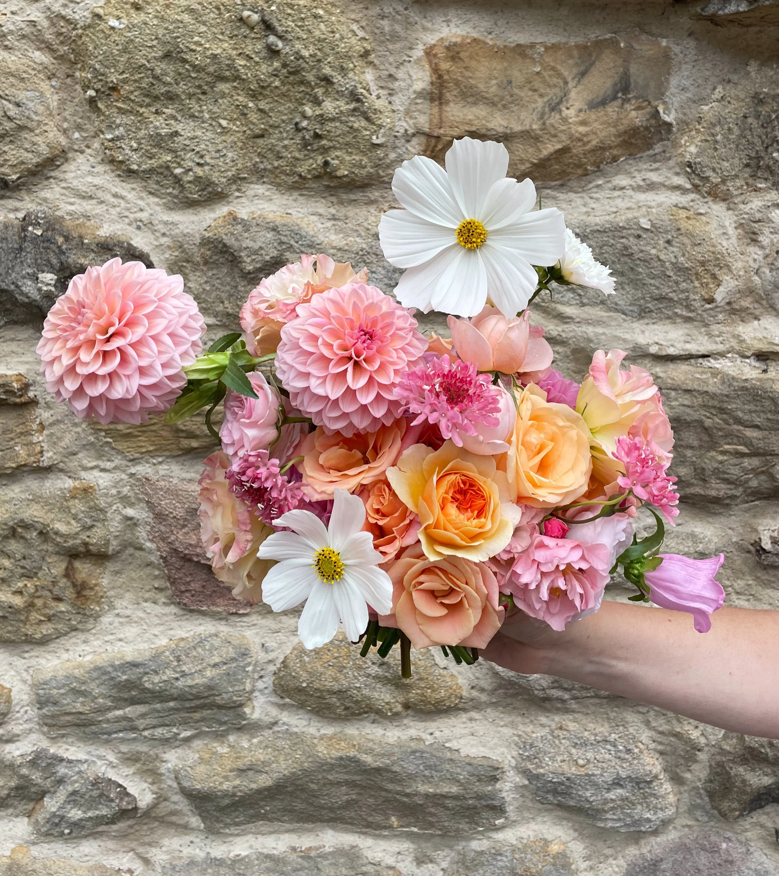 A hand holding a bouquet of various pastel-colored flowers against a stone wall background.