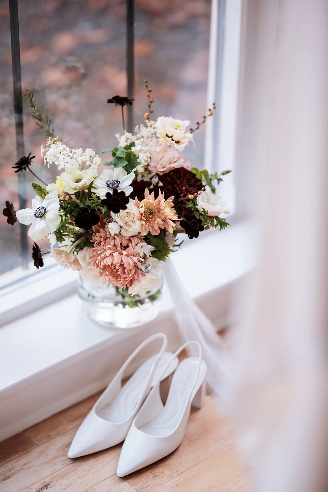 White high heel shoes placed on wooden floor near a window with a bouquet of pink, white, and dark-colored flowers in a glass vase.