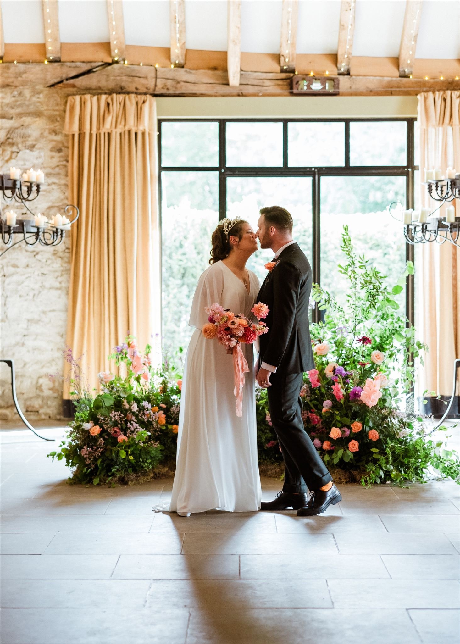 A bride and groom standing close together, about to kiss, in front of large windows with curtains. The setting features floral arrangements and warm lighting.