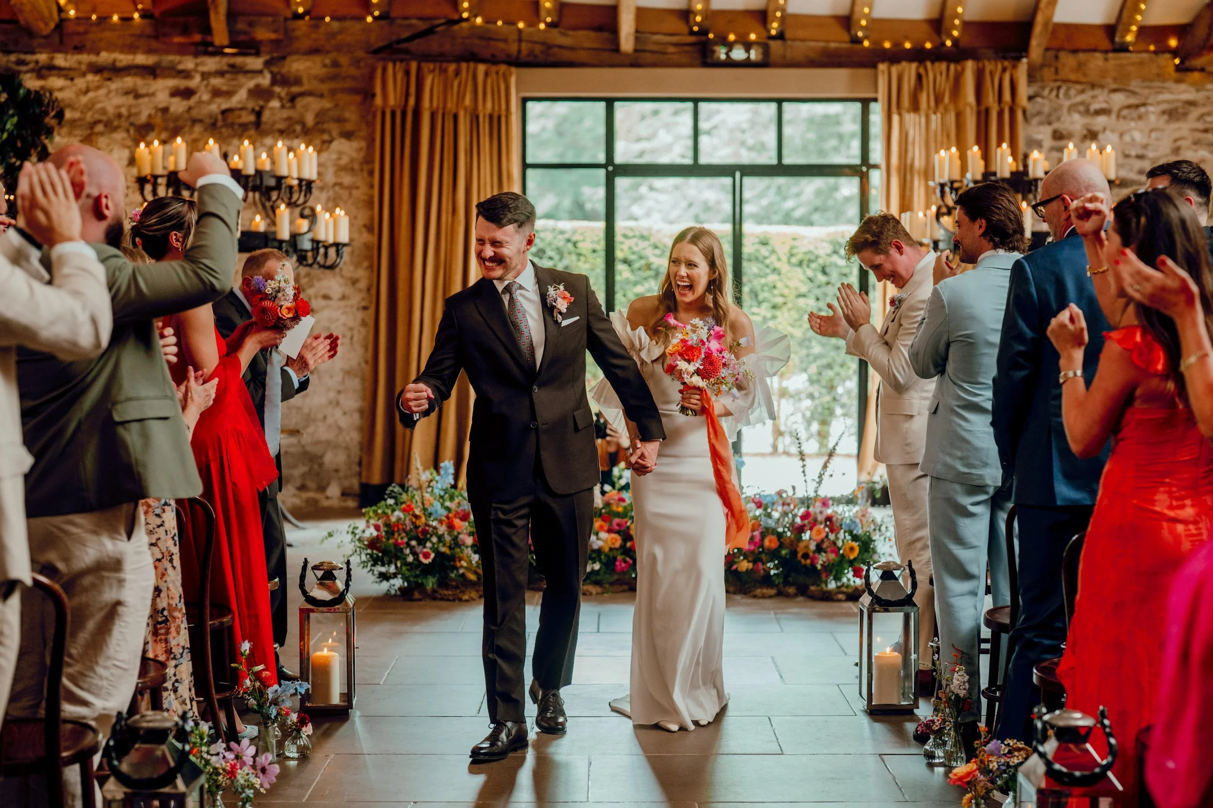 Just married couple walking back down the aisle at the Tithe Barn, Bolton Abbey, North Yorkshire. Bright and modern wedding flowers with meadow ceremony next and bud vases.