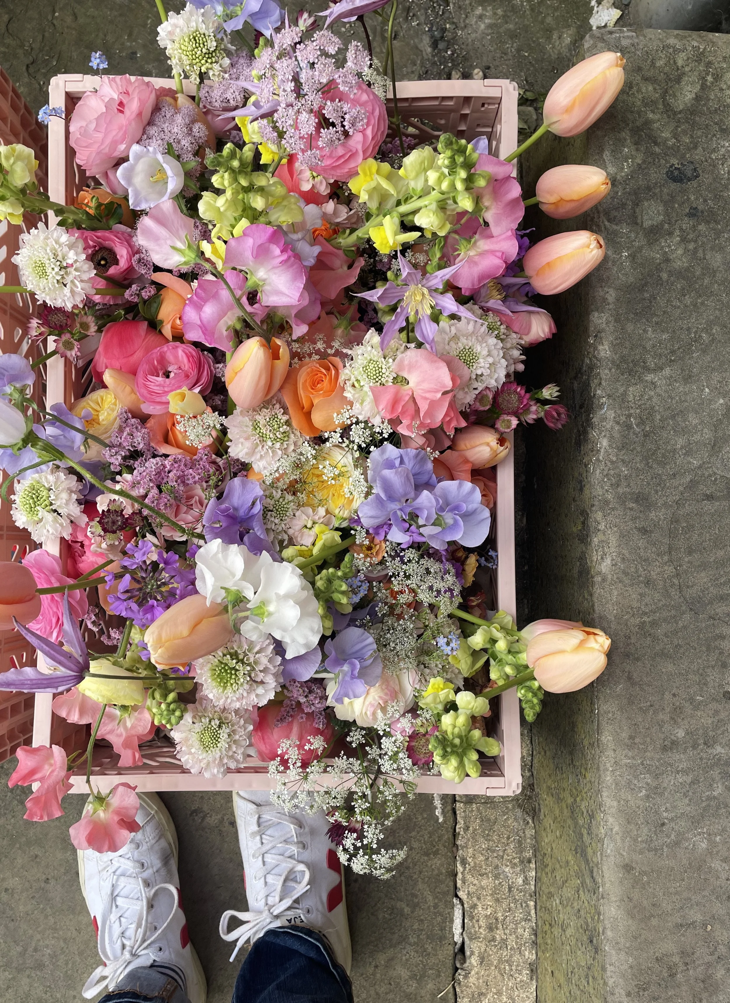A pastel pink basket filled with various colorful flowers including tulips, roses, lilies, and other small flowers, placed on a concrete ground.
