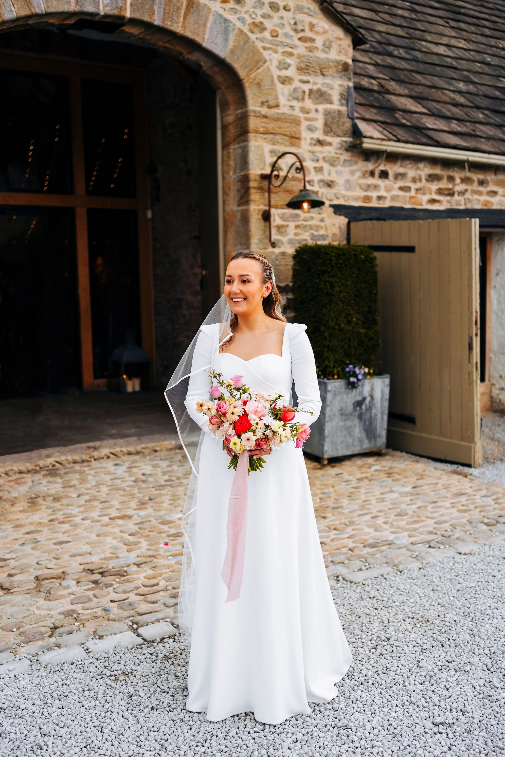 bride standing outside The Tithe Barn, Bolton Abbey, North Yorkshire holding her spring flowers bouquet with butterfly ranunculus, paper whites and tulips in pinks, whites and greens