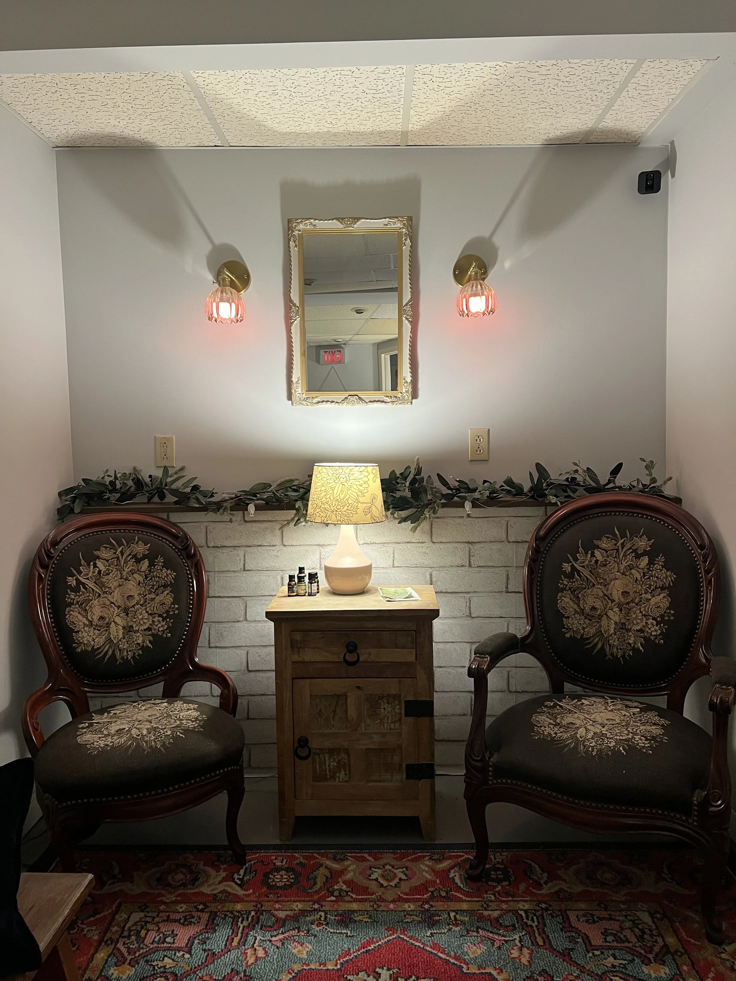 Vintage living room with two upholstered armchairs featuring floral embroidery, a small wooden side table with a table lamp, a mirror with gold frame, red wall sconces, greenery, and a patterned rug.