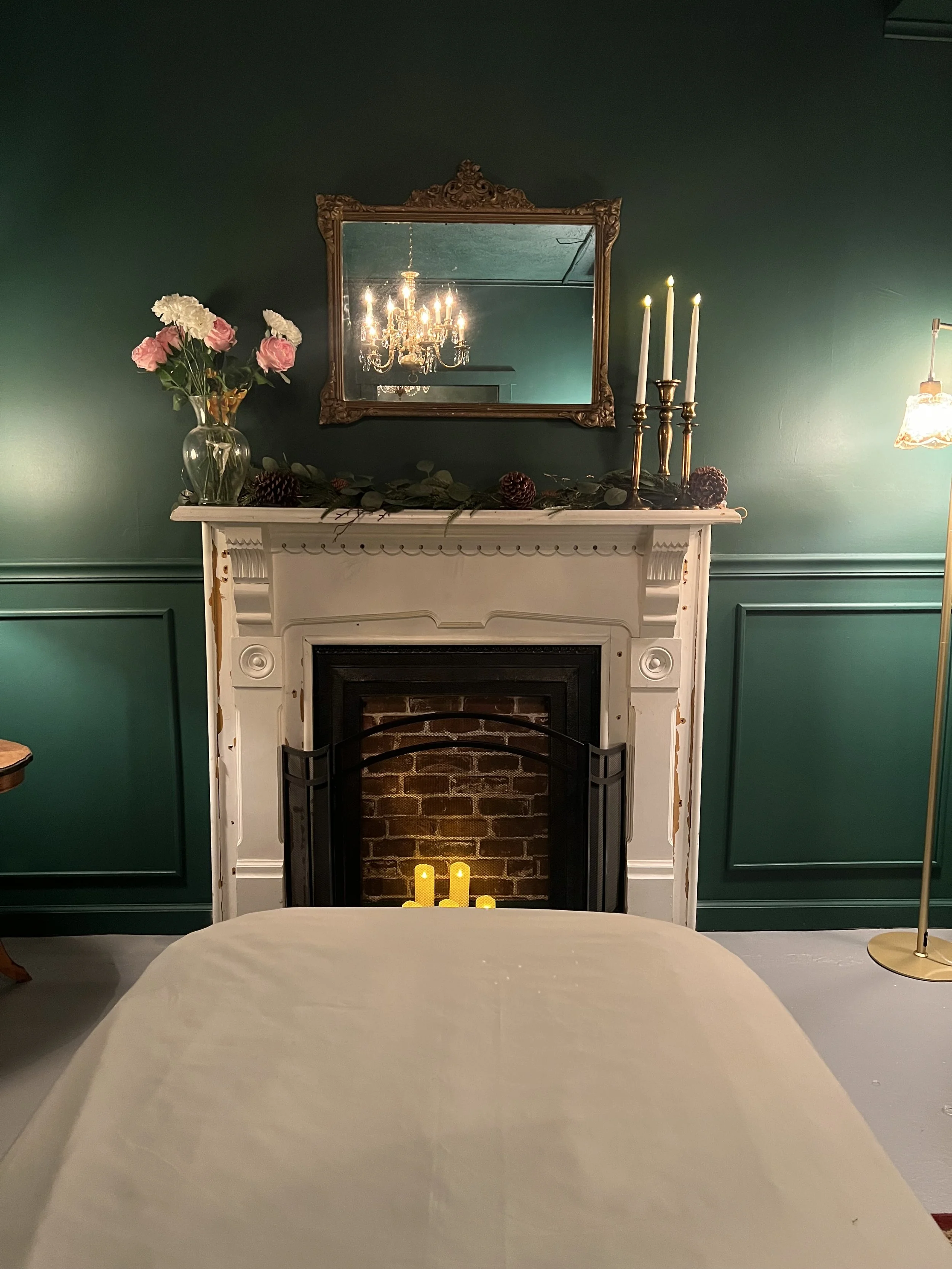 A living room with a white fireplace, decorated with a mirror, flowers, candles, and a chandelier reflection in the mirror.