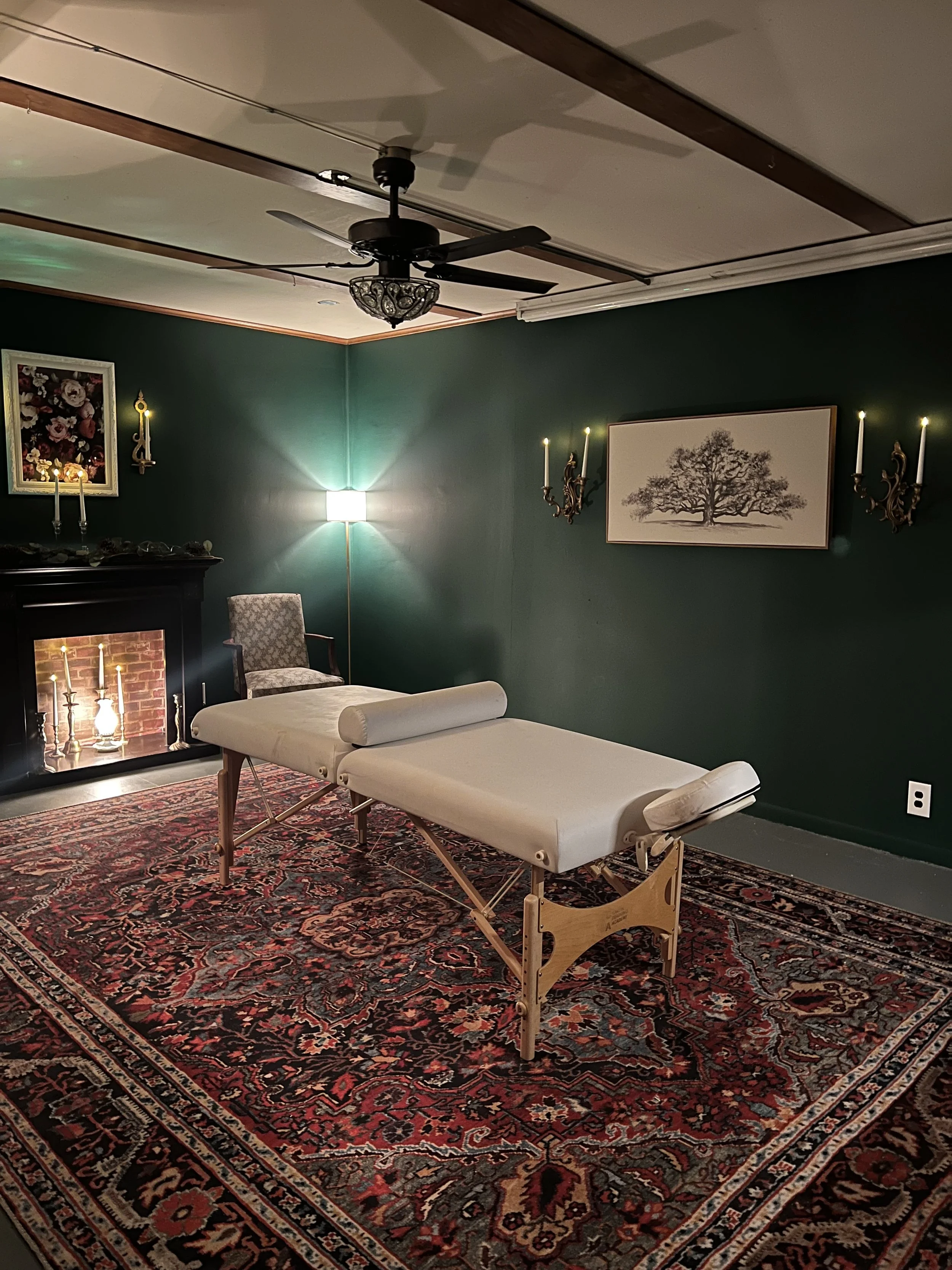 A massage therapy room with a massage table covered in white sheets, a patterned armchair beside it, a classic fireplace with candles, dark green walls decorated with framed artwork and wall-mounted candle holders, and a Persian rug on the floor.