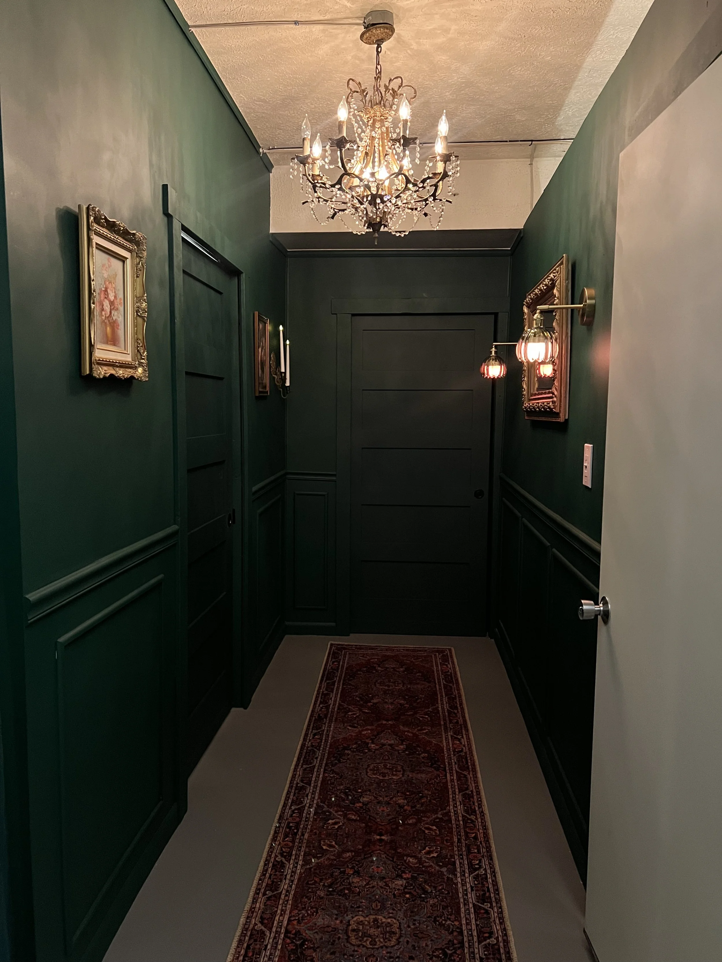 Decorative hallway with green walls, ornate paintings, a chandelier, wall sconce lights, and an oriental rug.