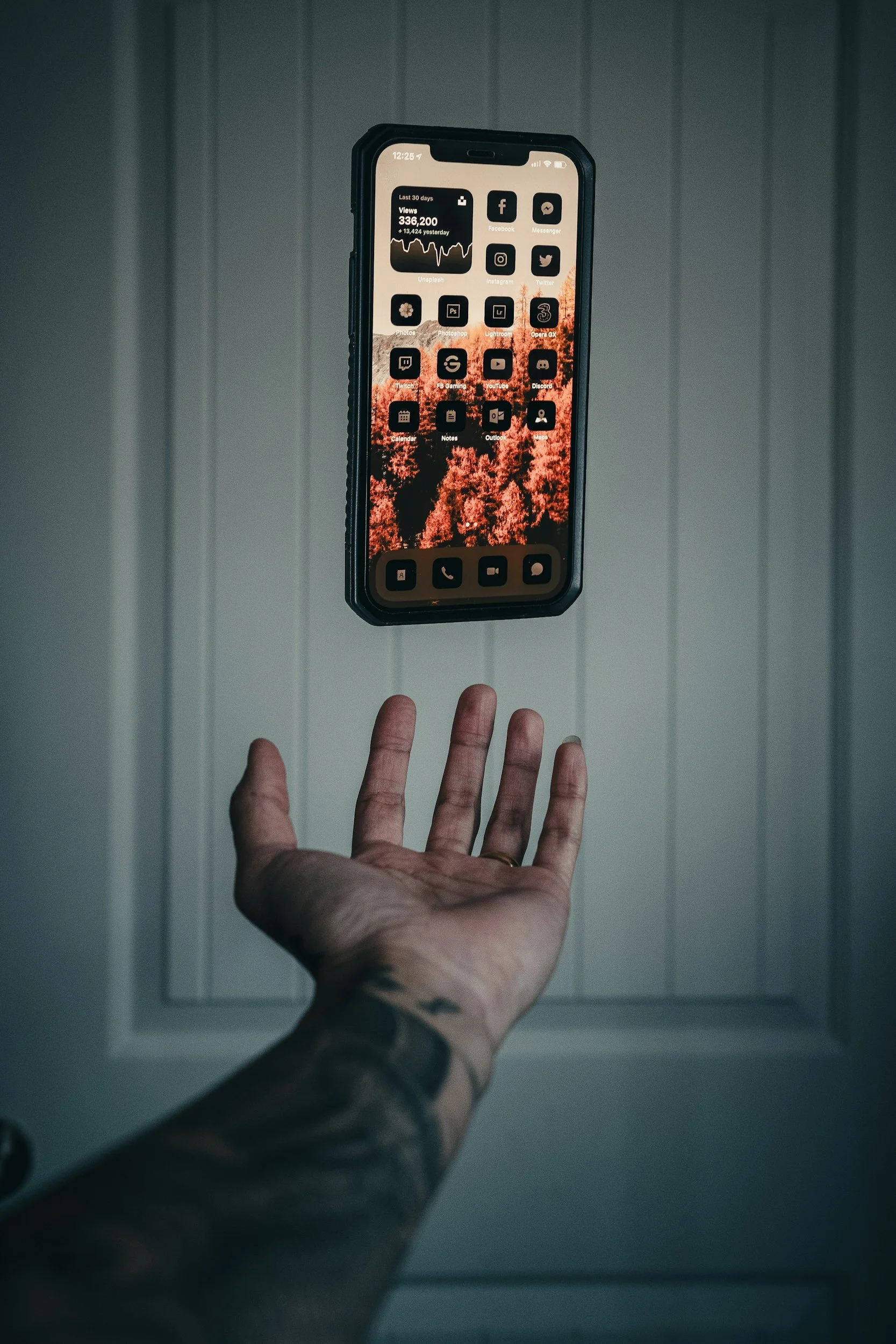 A smartphone floats above a tattooed hand. The screen displays a custom iOS interface with orange icons and a forest wallpaper.