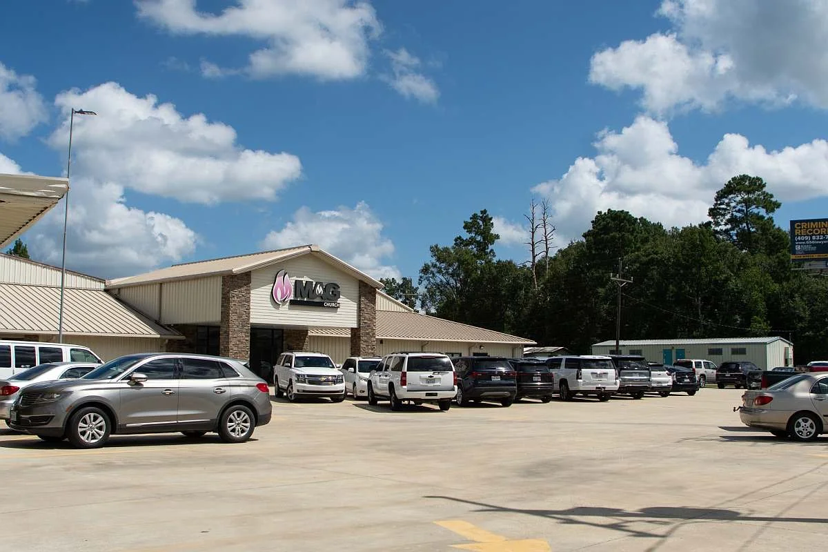 Front parking lot of MAG Church showing several parked cars on a Sunday morning under a partly cloudy sky.