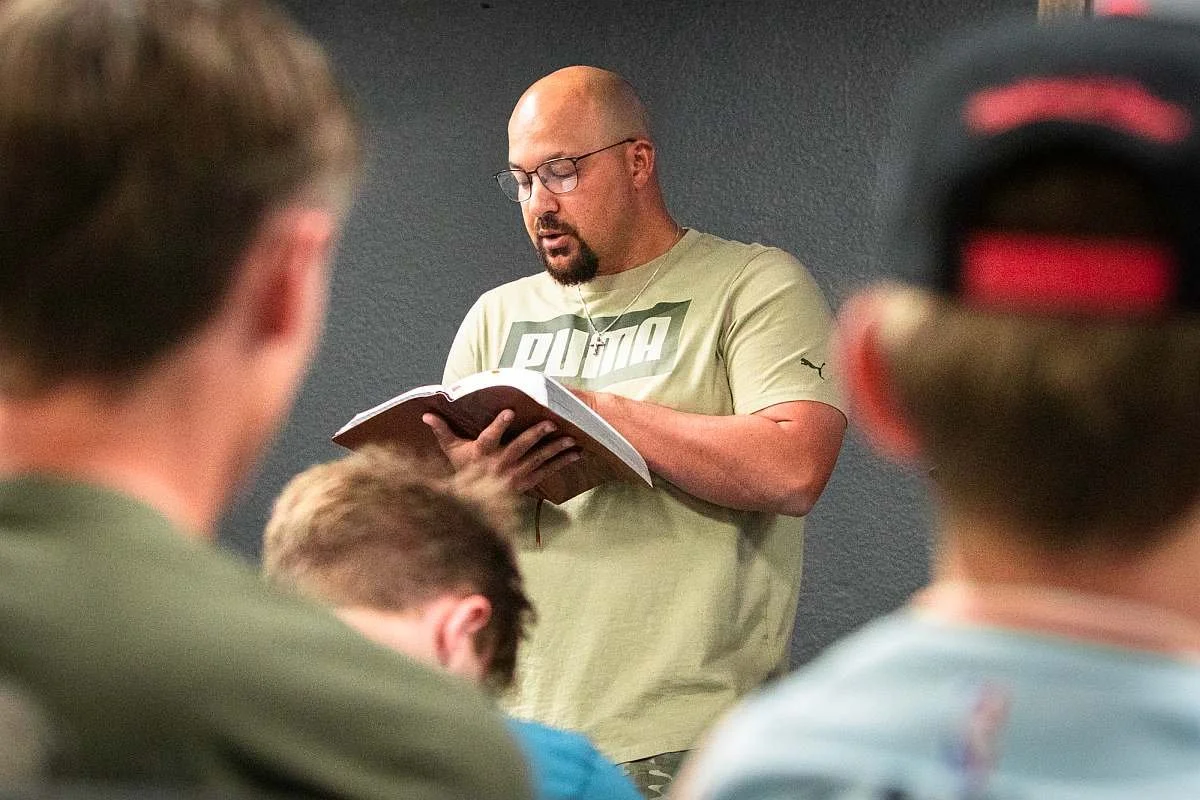 Pastor Mark Suggs, who is bald with glasses, is preaching a message from the Bible while standing in front of a dark wall, with several young people sitting in front of him, some with blurred heads and one wearing a cap.