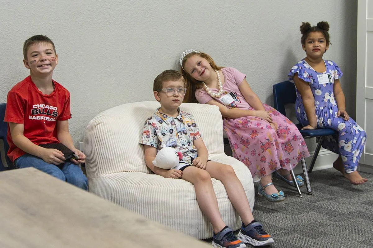 Four children sitting on chairs and a sofa against a plain wall, with various expressions. One boy holding a game controller, two girls dressed in colorful outfits, and a boy with glasses holding a plush toy.