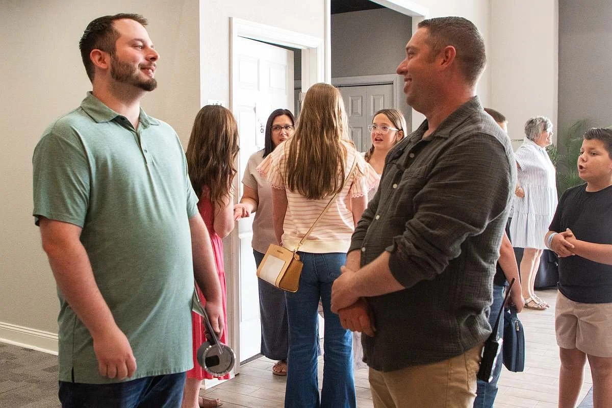 Two men are talking and smiling in the foreground in the MAG Church foyer with other people in the background talking as well.
