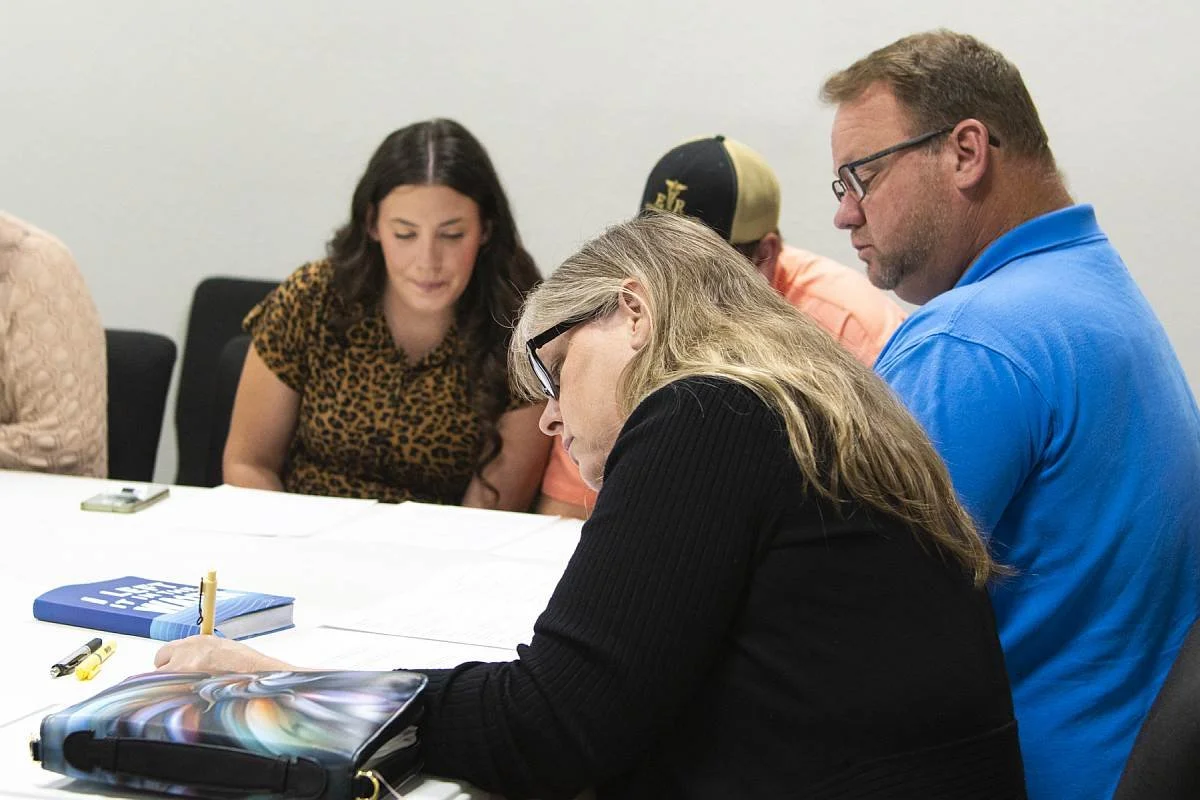 Four people sitting around a table engaged in a discussion or working, with notebooks, a Bible, pens, and a phone on the table.