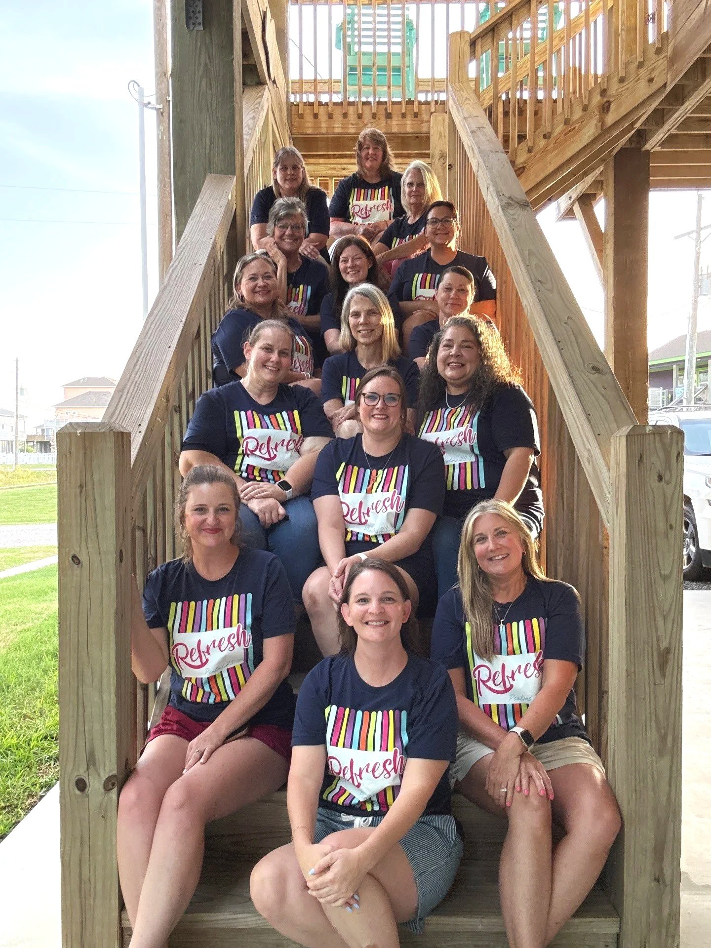 Group of women sitting on wooden stairs outdoors, beside a beach house at Crystal Beach, TX, smiling, wearing matching black T-shirts with colorful design and the word 'Refresh' on them.