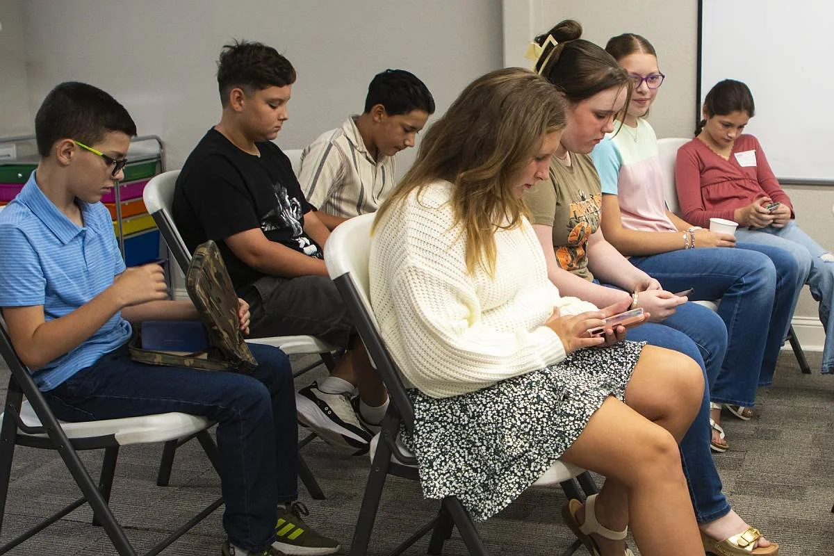 Group of children and young teens sitting in chairs, looking at a Bible app on their phones in a classroom.