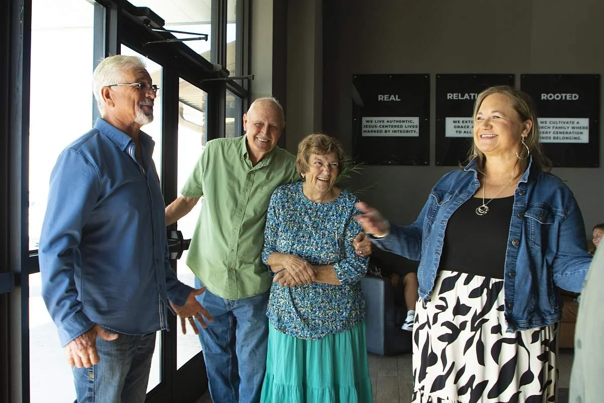 Four people, three men and one woman, are standing together and smiling inside the foyer of MAG Church near large windows, having a conversation.