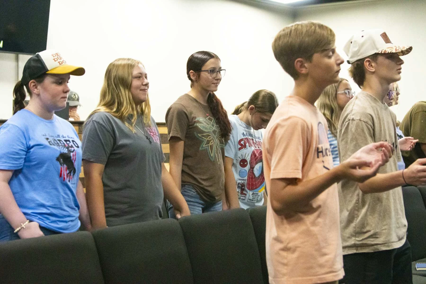 A group of young people standing in the MAG Church youth hall, with most of them holding their hands in a prayer-like position and their eyes closed in a moment of prayer.