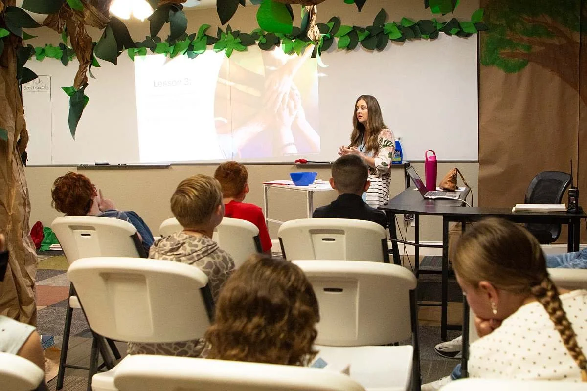 A classroom with a teacher standing in front of students, delivering a lesson. The students are seated in white chairs facing the teacher, who is standing near a whiteboard with a projected slide. The classroom is decorated with paper leaves and trees, creating a jungle or forest theme.