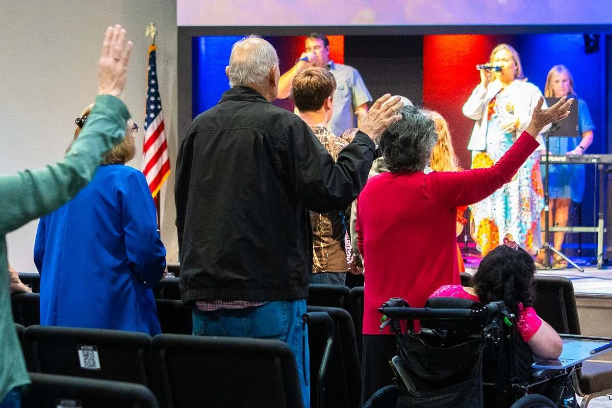 People standing and raising their hands during a worship service at MAG Church, with a worship team playing and singing on a platform in the background.