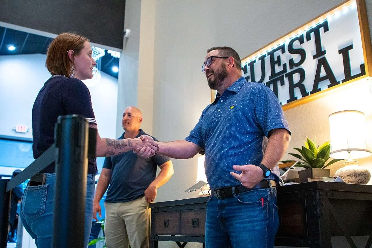 A man and woman shaking hands indoors at MAG Church's guest central area, with another man standing in the background near a desk and a sign that says 'GUEST CENTRAL'.