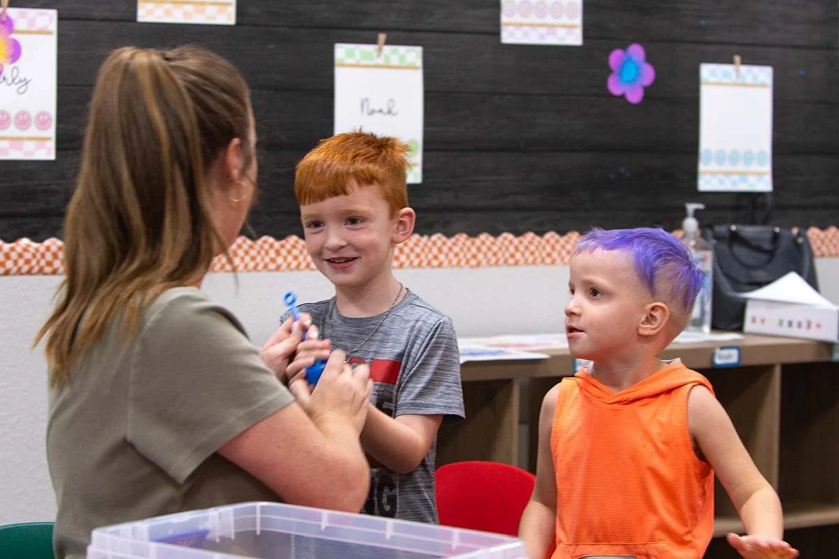 A teacher with brown hair interacts with two young boys, one with red hair and the other with purple hair, in a preschool classroom setting.