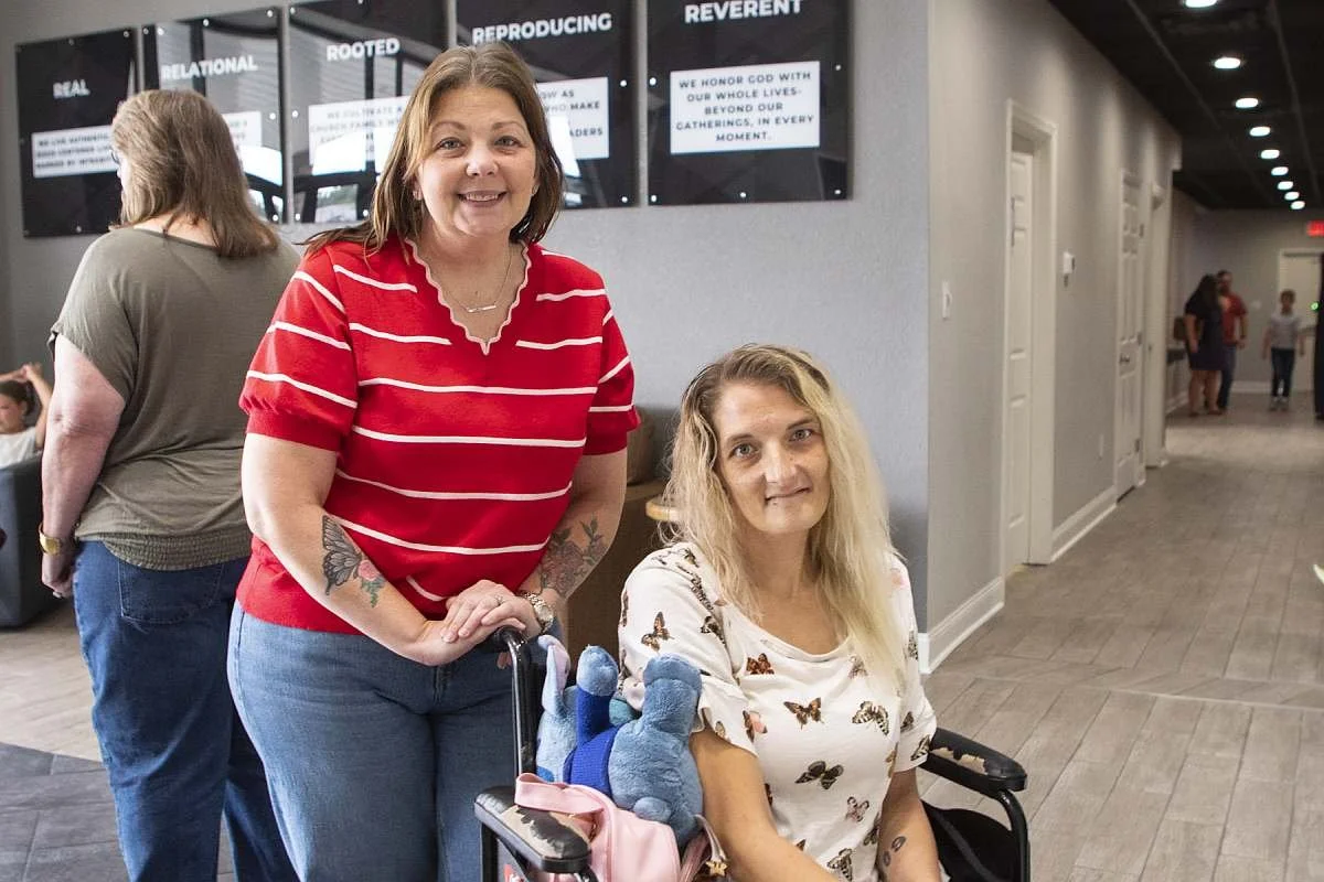 Two women in the MAG Church lobby, one standing and smiling, the other sitting in a wheelchair with a stuffed animal, both looking at the camera. There are other people in the background and signs on the wall behind them.