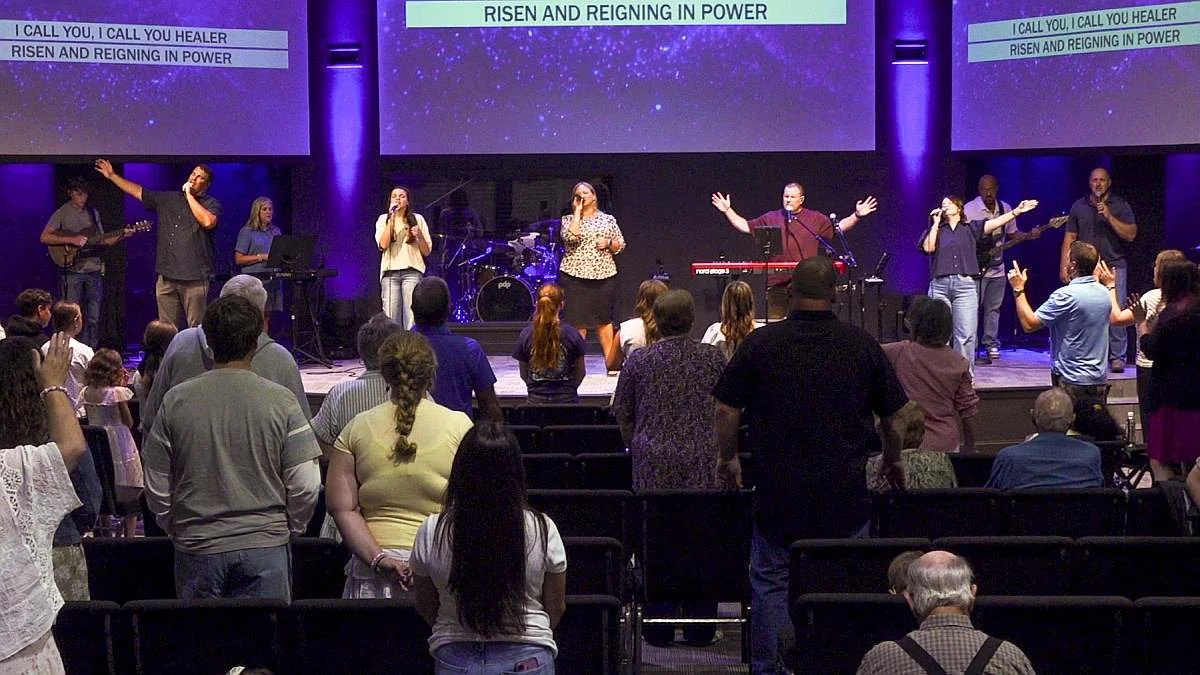 A group of people worshiping and singing on a stage during a church service, with a congregation standing and raising their hands in front of them. The stage has a band and large screens displaying lyrics that say "I CALL YOU, I CALL YOU HEALER" and "RISEN AND REIGNING IN POWER."