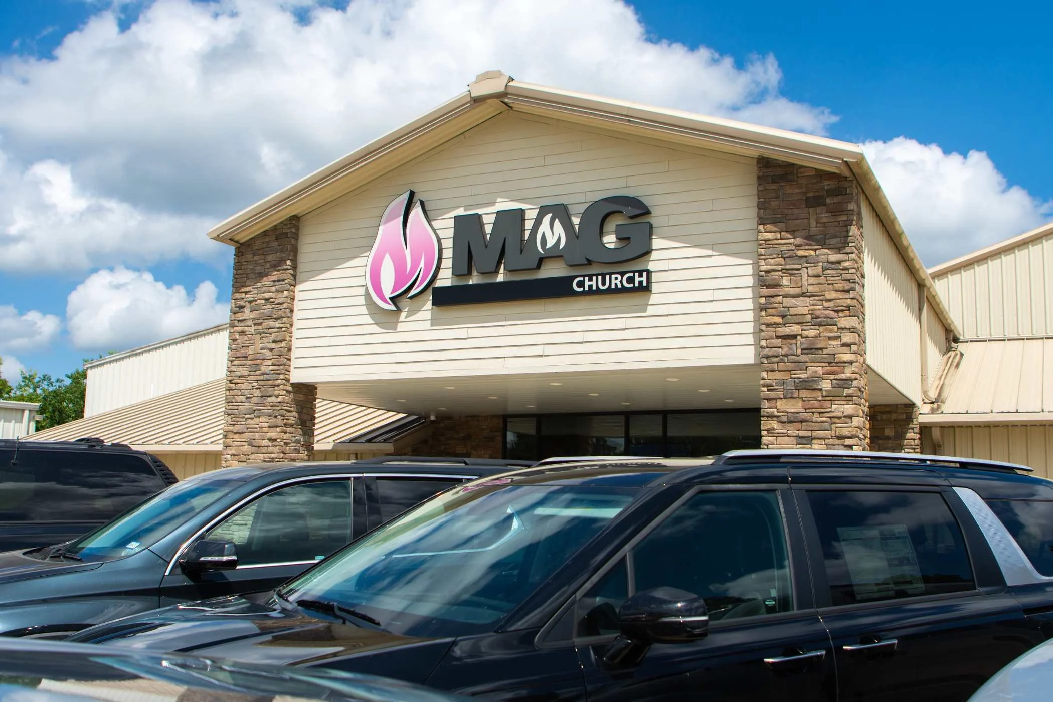 The exterior of a church building with a sign that reads 'MAG Church' and a stylized flame logo. There are parked cars in the foreground, and the sky is partly cloudy.
