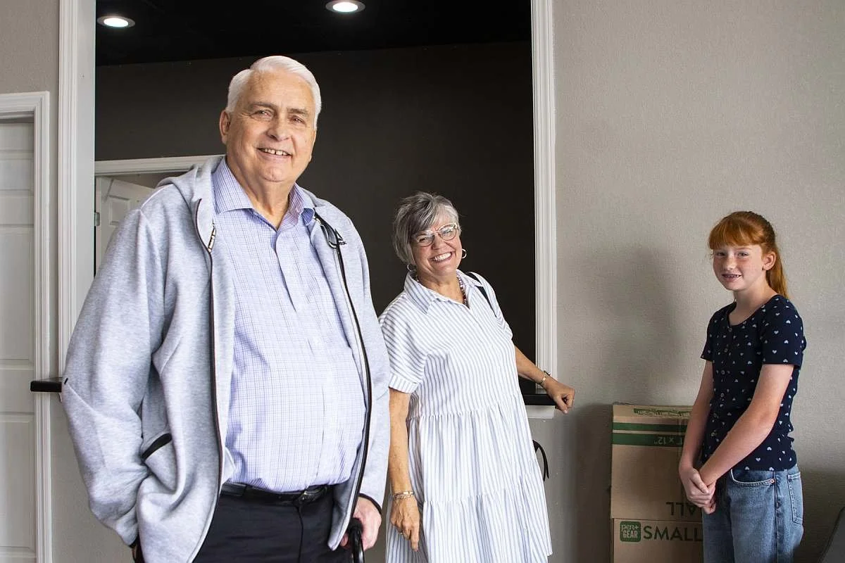 An elderly man and woman standing indoors with a young girl, smiling at the camera. The man is wearing a light blue plaid shirt and a gray zip-up hoodie, holding a cane. The woman is wearing glasses and a white striped dress. The girl is wearing a dark blue shirt with white polka dots and jeans.