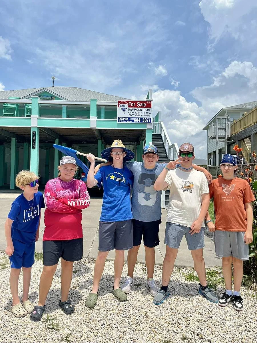 Group of six kids from MAG Church  posing outdoors in front of a house with a 'For Sale' sign, dressed in summer clothes, some wearing sunglasses and hats, one holding a blue stick or flag, against a partly cloudy sky.