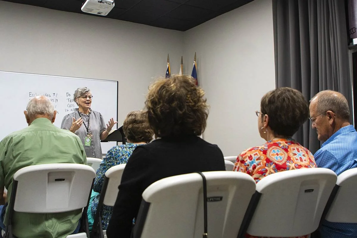 A woman standing at the front of a room giving a presentation to an audience of several people sitting in white chairs, with a whiteboard behind her bearing handwritten Bible verses..