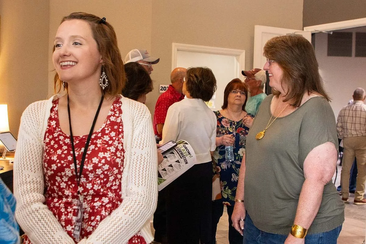 Two women smiling and engaging in conversation inside the MAG Church foyer, with several other people chatting in the background.