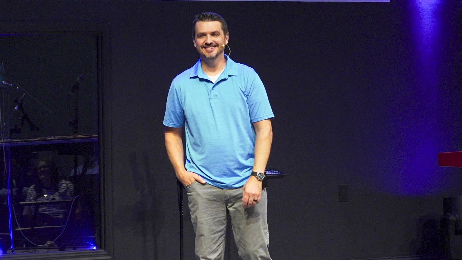 A man with dark hair and a beard wearing a blue polo shirt and gray pants, standing against a dark background, smiling and wearing a microphone headset.