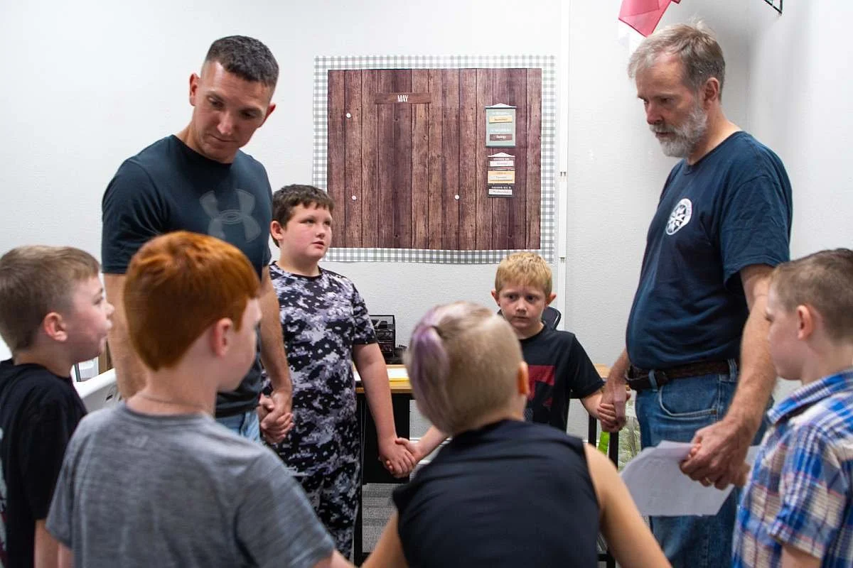 A group of young boys and two adult males holding hands in a circle, praying in a classroom during their Royal Ranger's class.