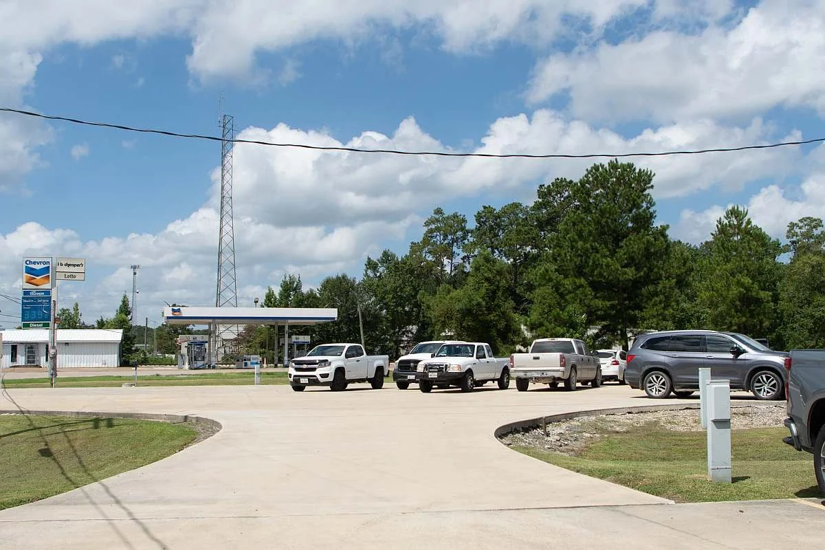 Side parking lot of MAG Church, with gas station Chevron and Texaco signs in the background. There are several parked cars including a white truck, trees, blue sky with clouds, and a curved concrete driveway.