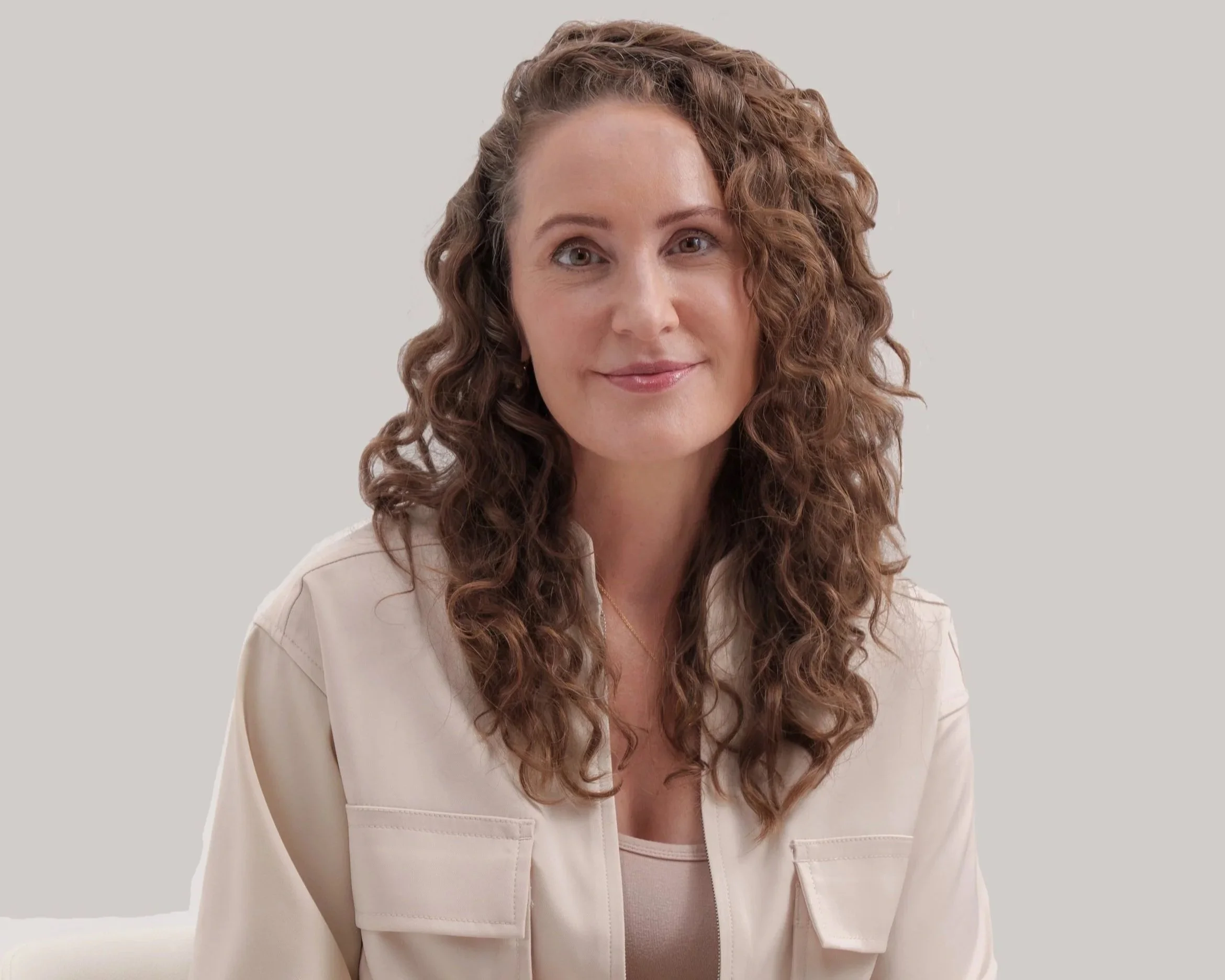 A woman with curly light brown hair and light skin, wearing a light-colored blazer and a beige top, smiling softly against a neutral gray background.
