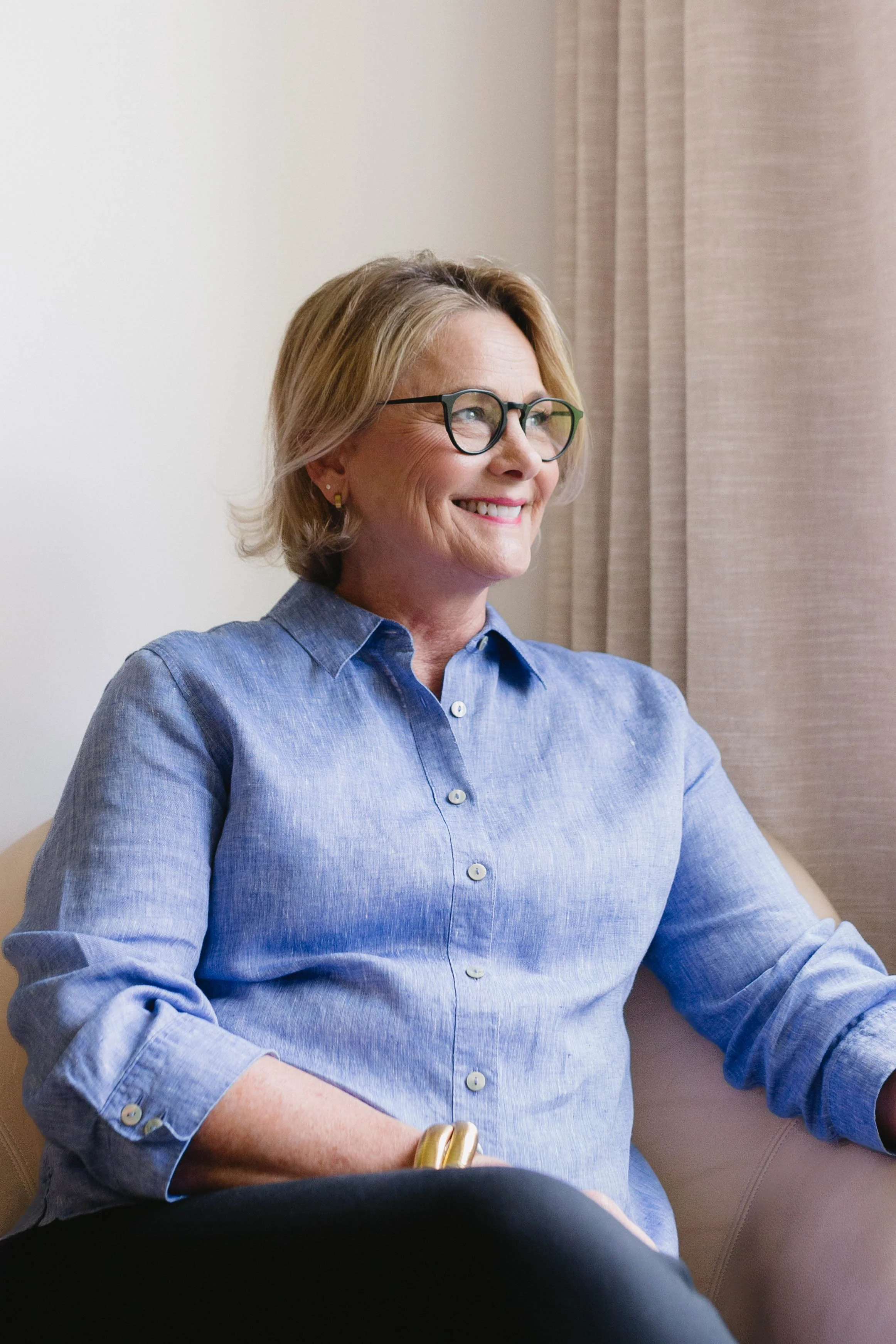 A middle-aged woman with short blonde hair, wearing glasses and a blue button-up shirt, smiling and sitting on a beige couch beside beige curtains.