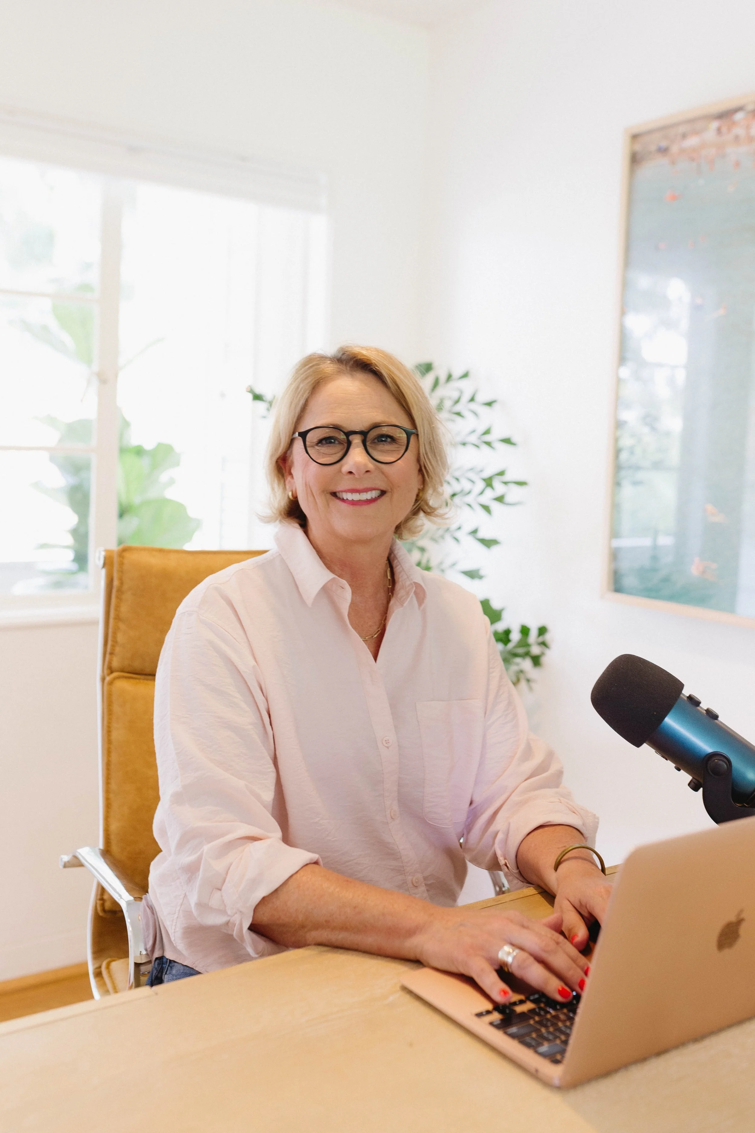 A woman with blonde hair, glasses, and a pink shirt sitting at a desk with a laptop and microphone, smiling at the camera in a bright room.