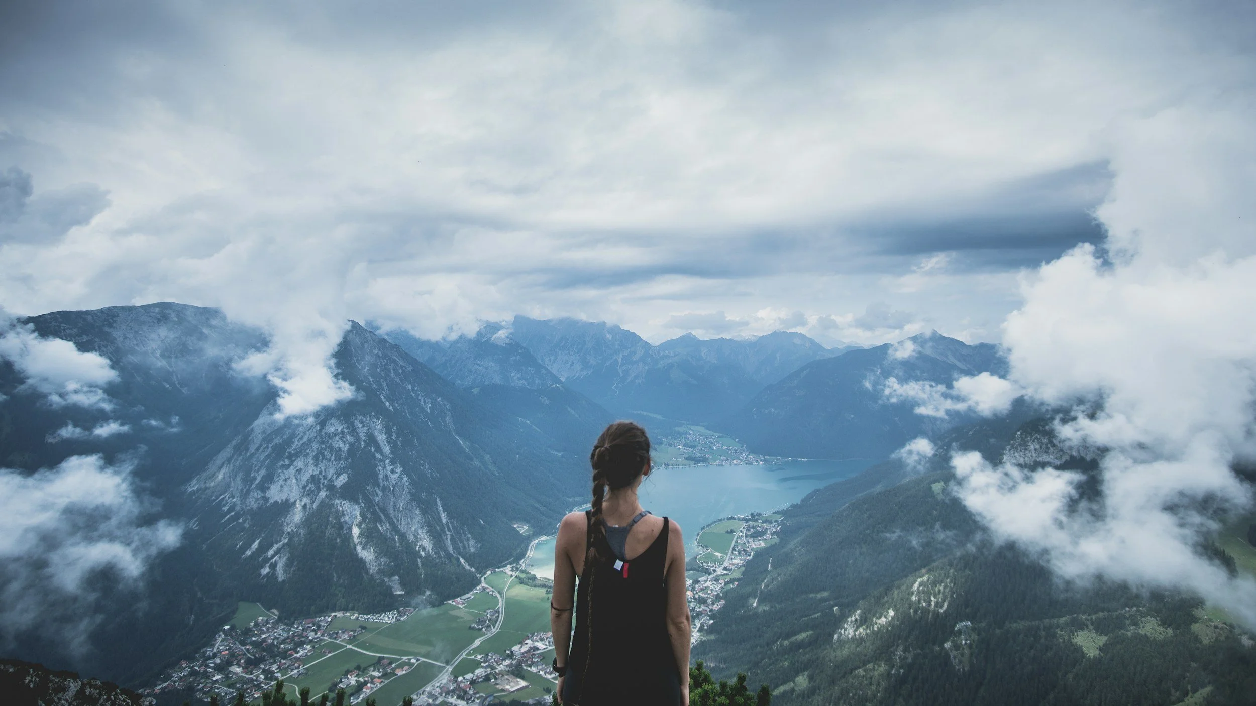 Person standing on a mountain ridge, gazing over a vast landscape of mountains and water, evoking reflection, growth, and possibility.