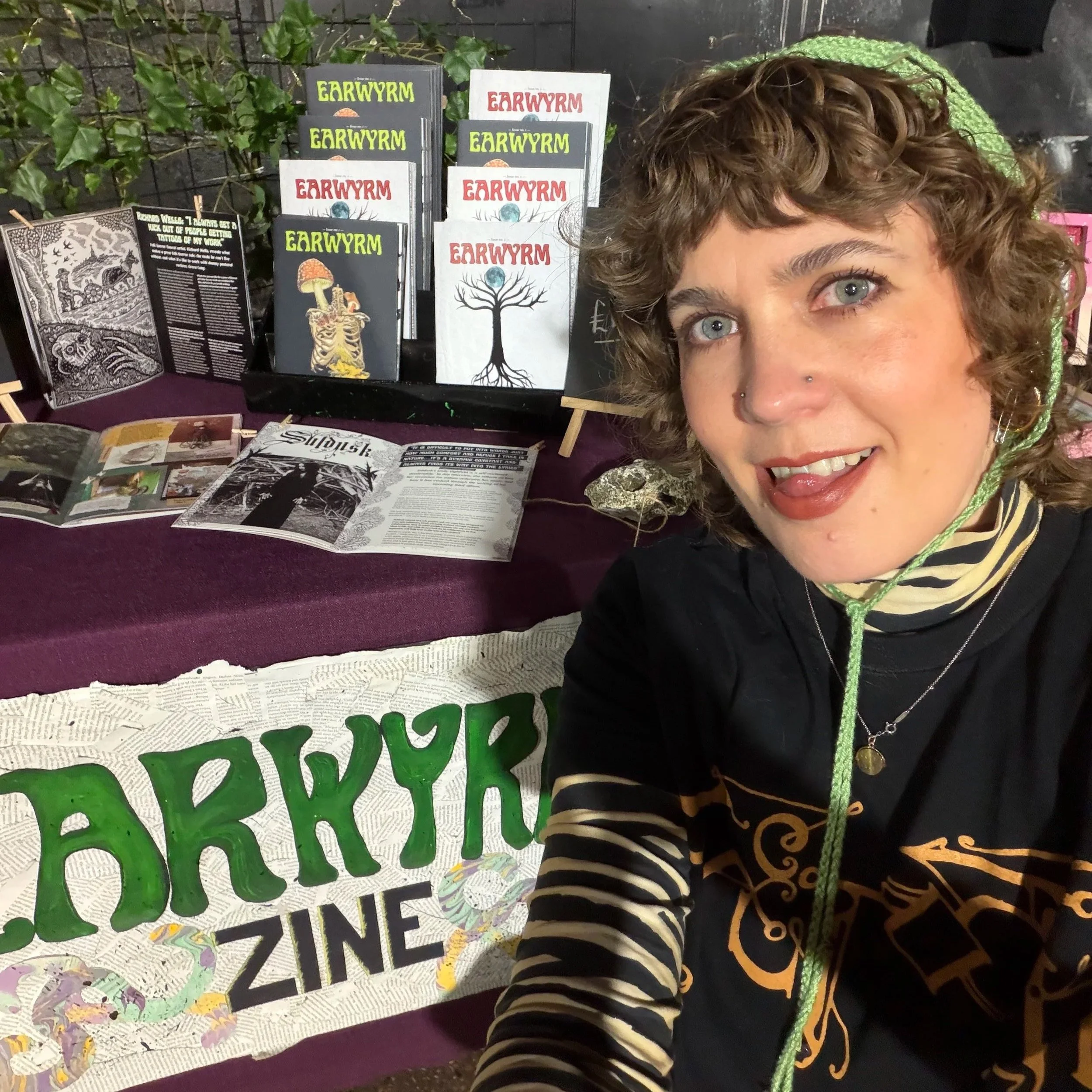 Me, a white millennial woman with curly light brown hair and blue eyes, kneeling in front of a market stall with a handmade collage sign reading Earwyrm Zine. There are print copies of my zine on a stand and laying out on the table.