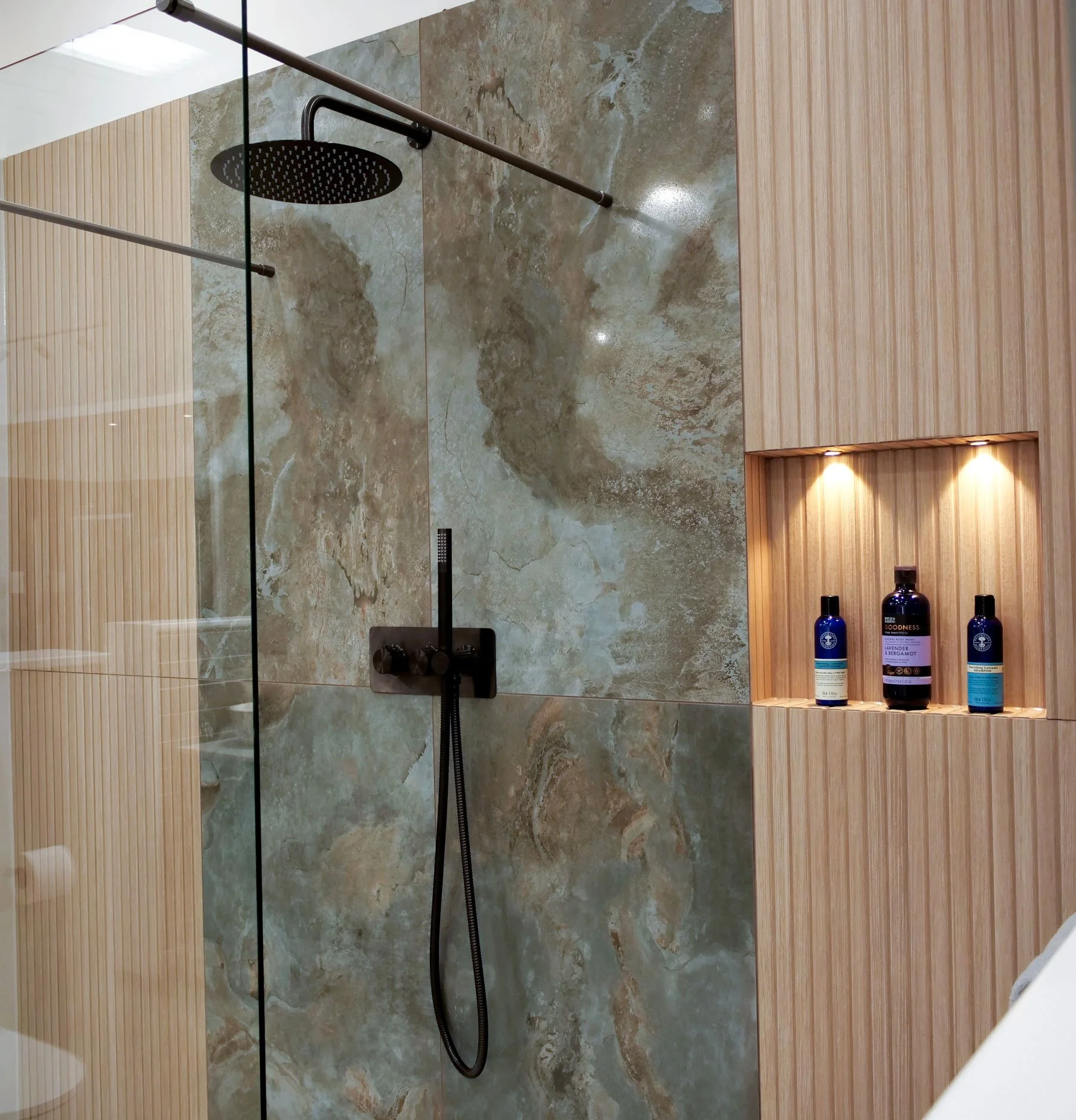 Modern shower area with large beige and brown marble tiles, black showerhead, and a built-in wooden shelf with three bottles of toiletries, illuminated by two small integrated lights.